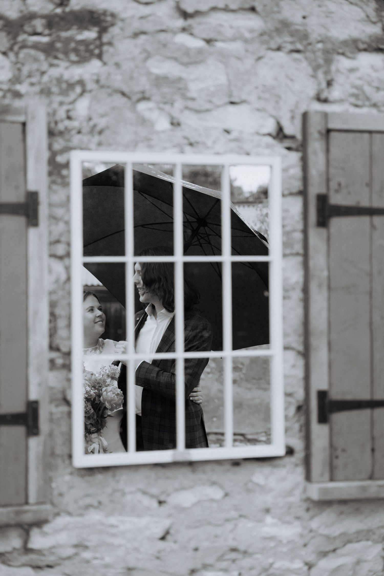 A bride and groom reflected in a window holding an umbrella on a rainy wedding day. by Braytmedia