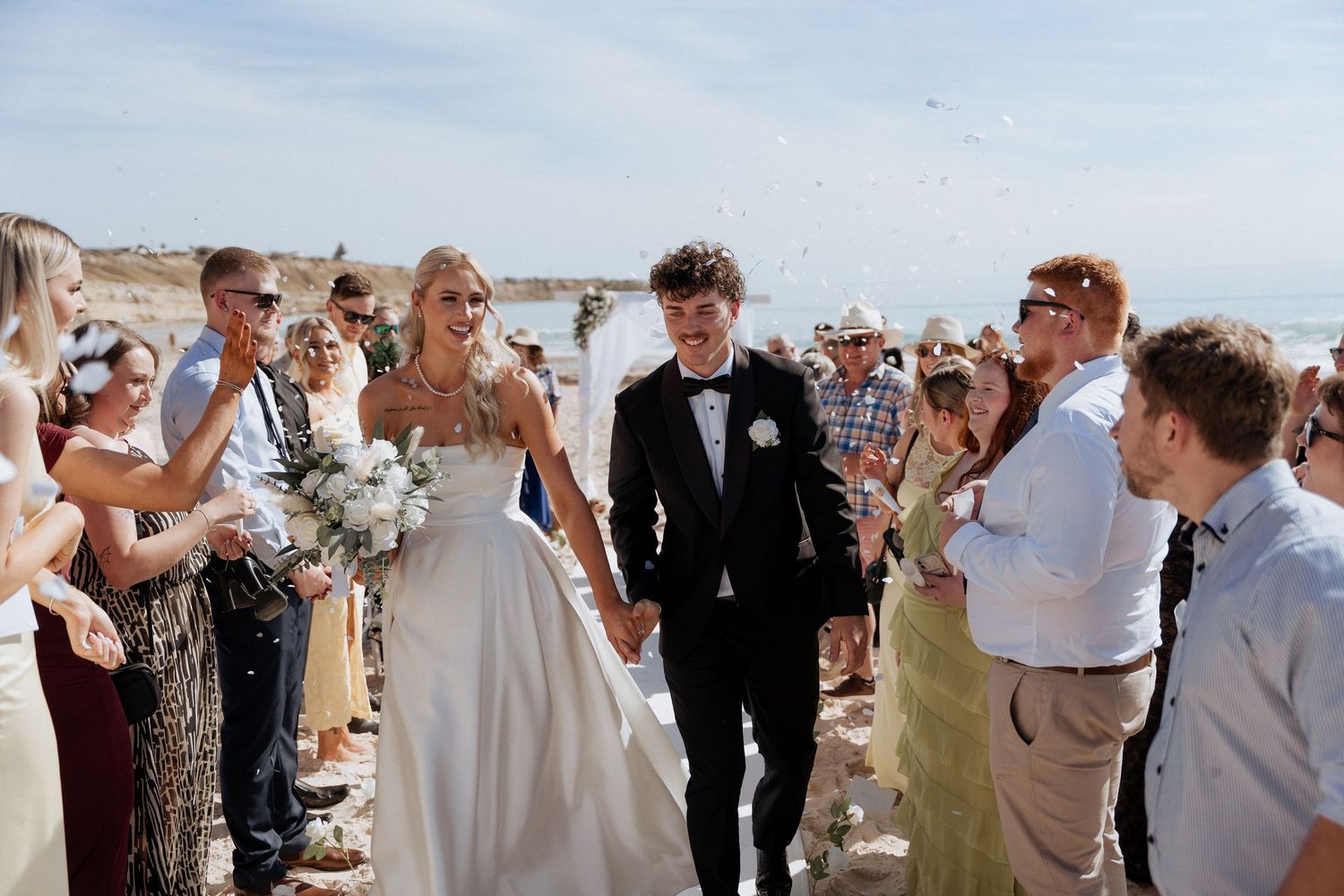 A joyful wedding celebration on the beach with guests throwing confetti as the newlyweds walk together.