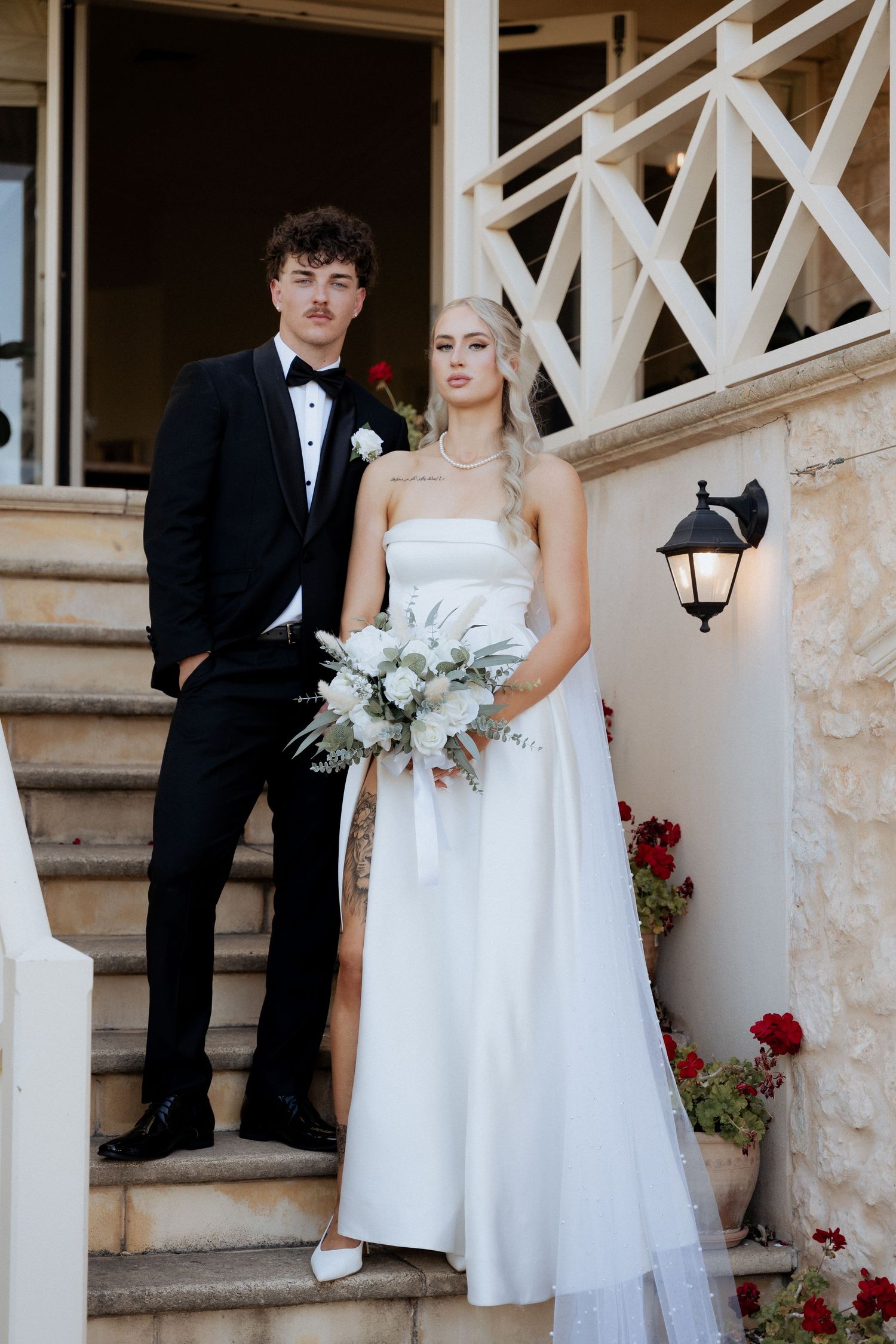 Wedding couple sitting on stone steps with white columns and red flowers in a romantic outdoor setting by braytmedia at the alton house