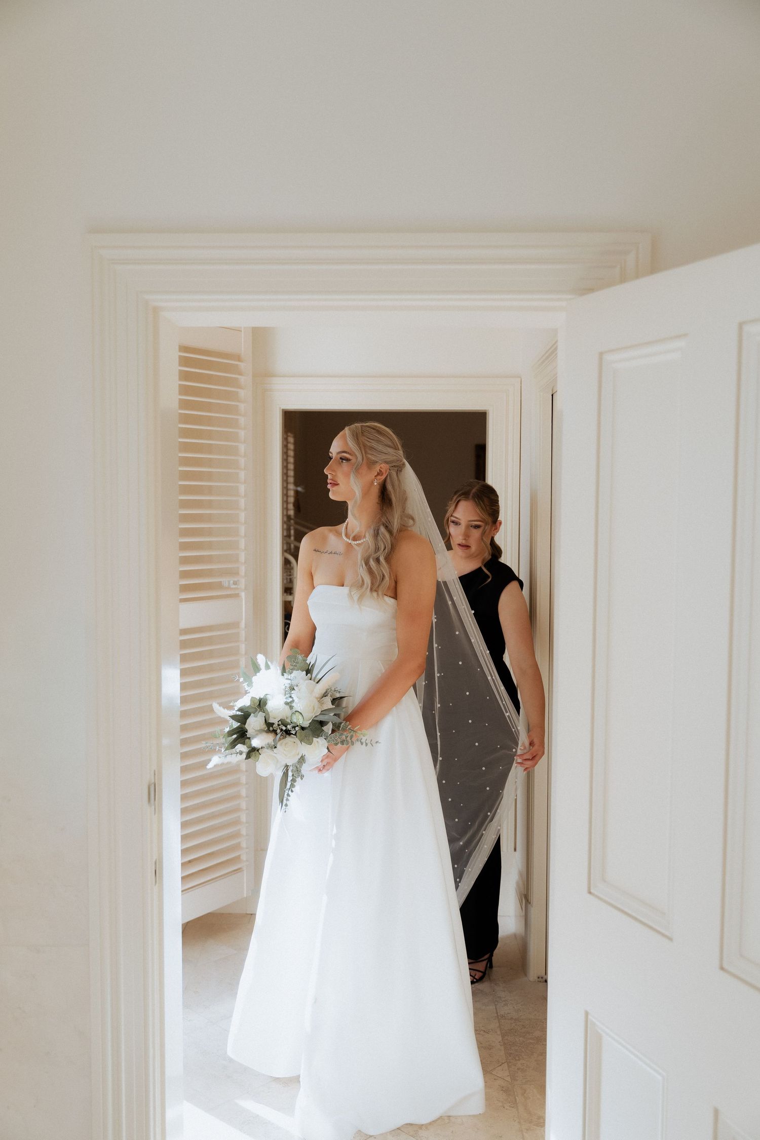 A bride in a flowing white wedding gown walks through a bright hallway while getting ready for her ceremony.