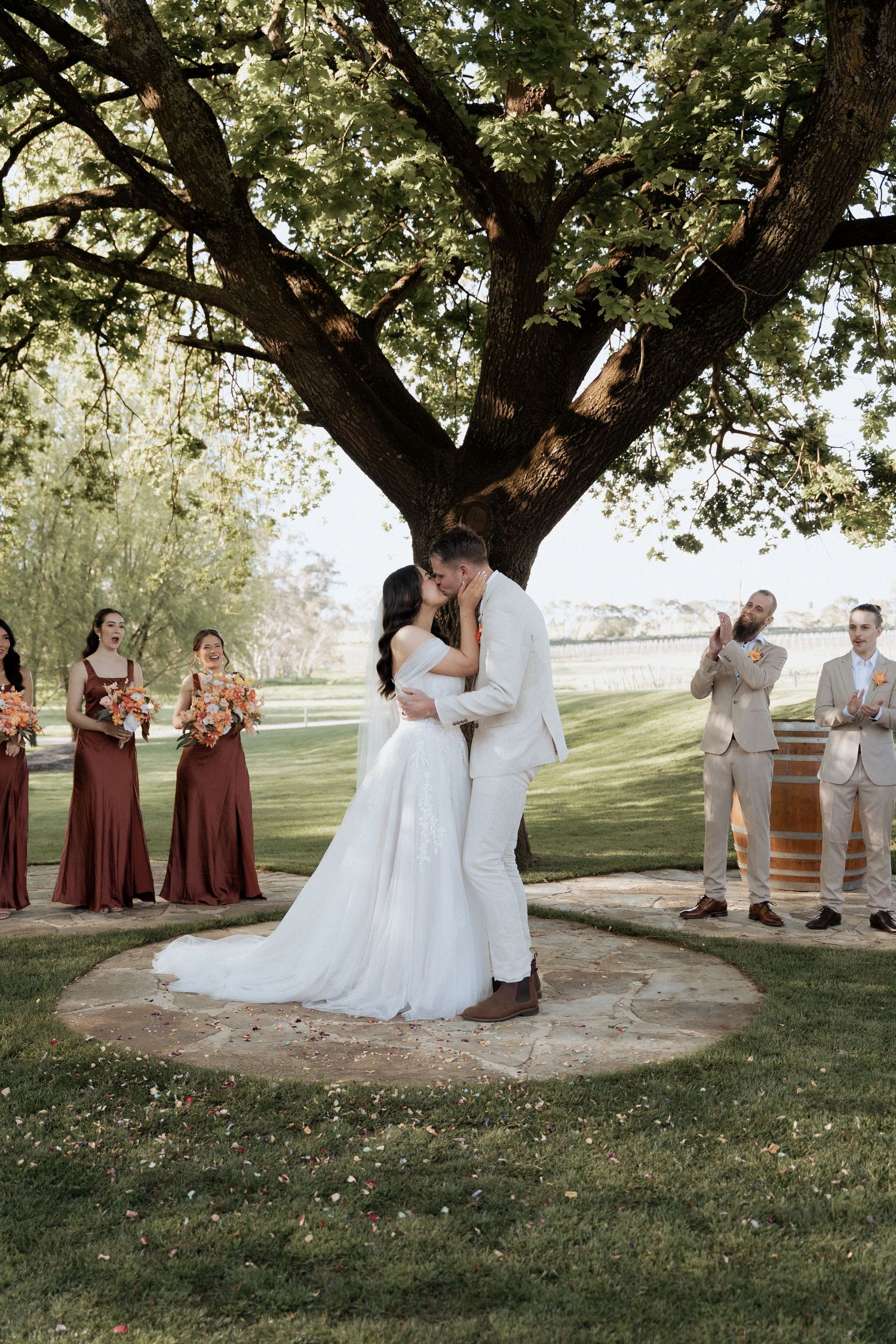 Wedding party poses under a shady tree with bridesmaids in burgundy dresses and groomsmen in light suits.