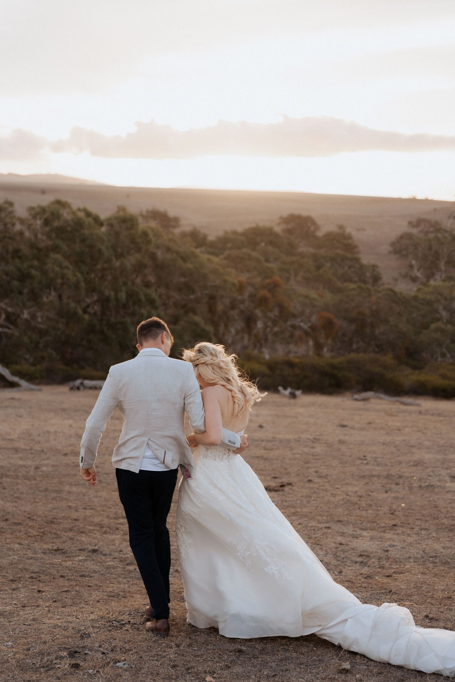 Bride and groom walk hand in hand across a hilltop during sunset with mountains in background.