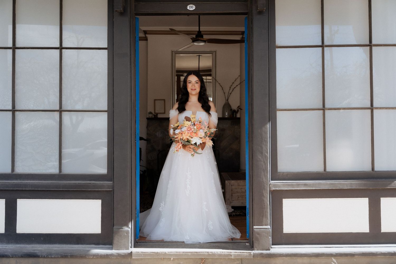 A bride in a white ballgown wedding dress holds a bouquet while standing in a doorway with frosted glass panels.