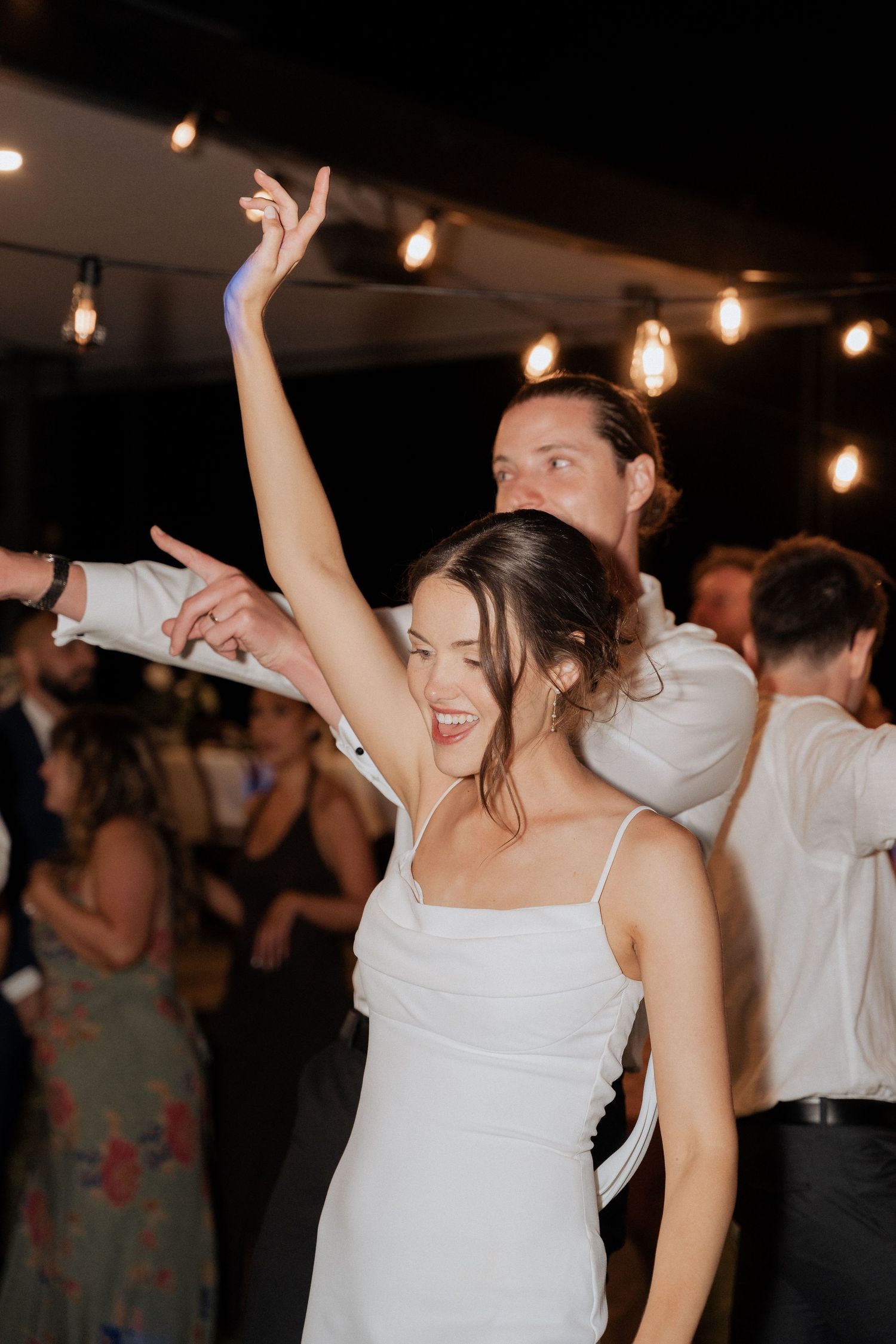 Wedding party guests dancing joyfully under string lights at an evening reception celebration.