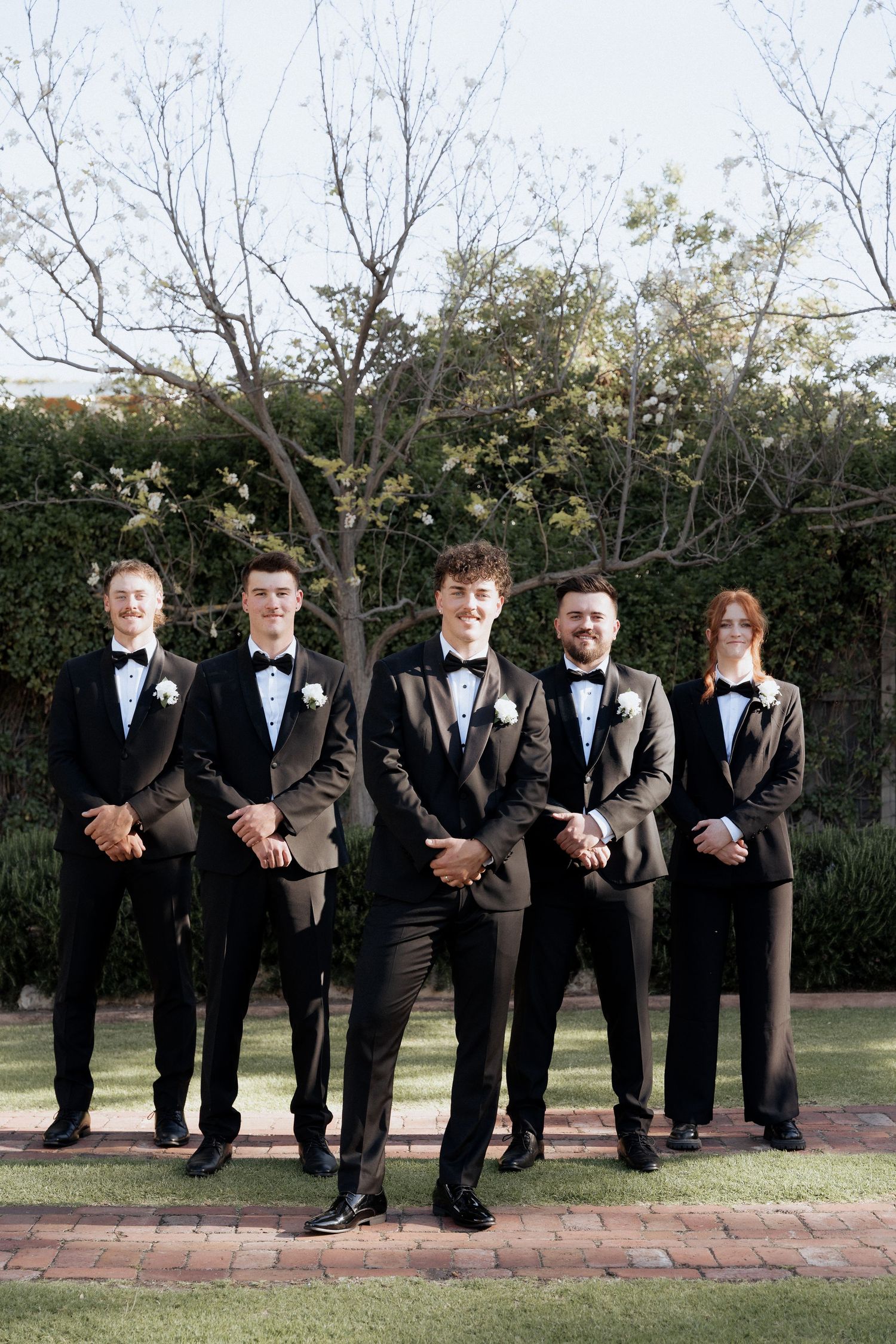 A wedding party in black tuxedos poses in a creative sequence along a brick path outdoors.