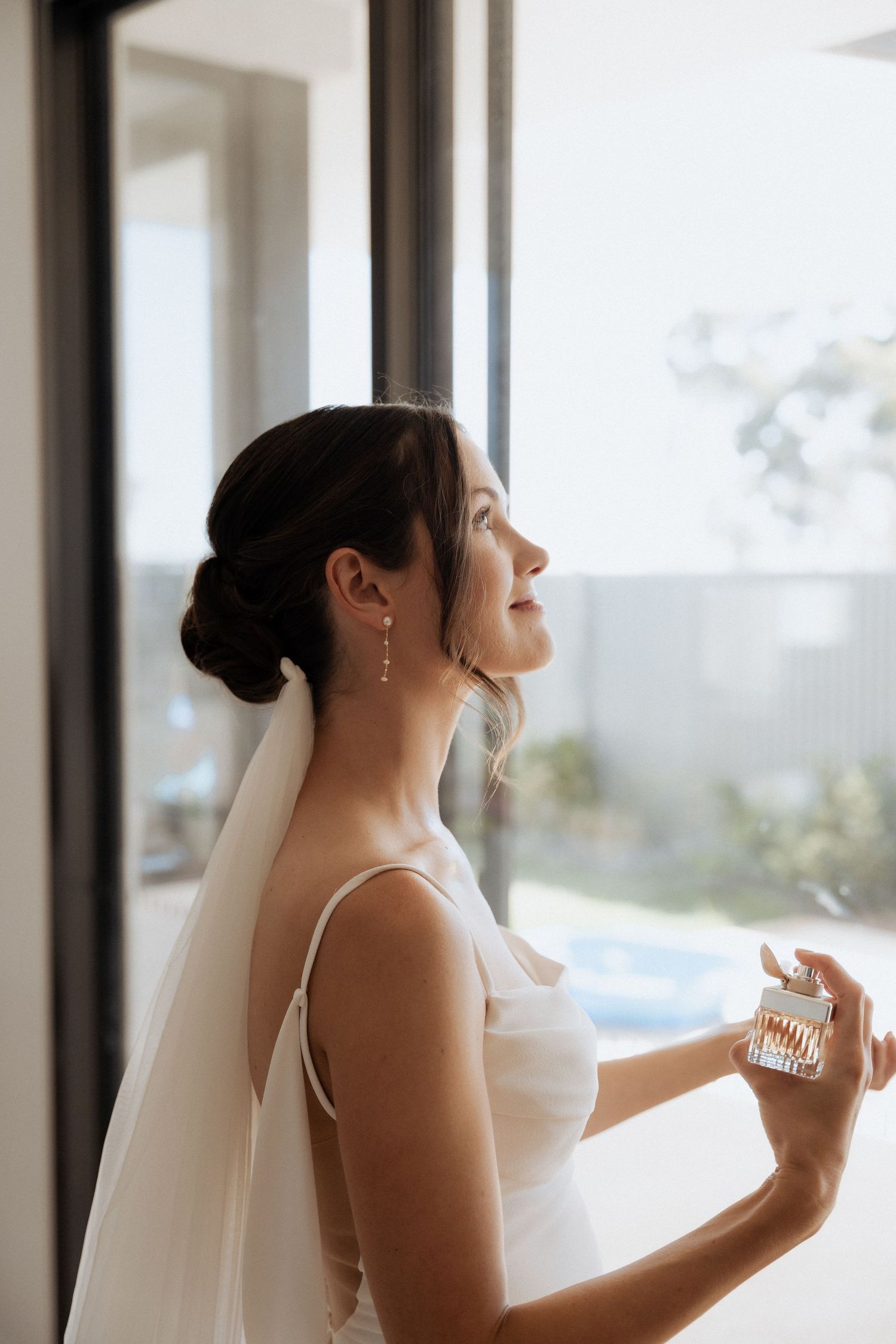 Bride in veil looking out large windows during wedding preparations.