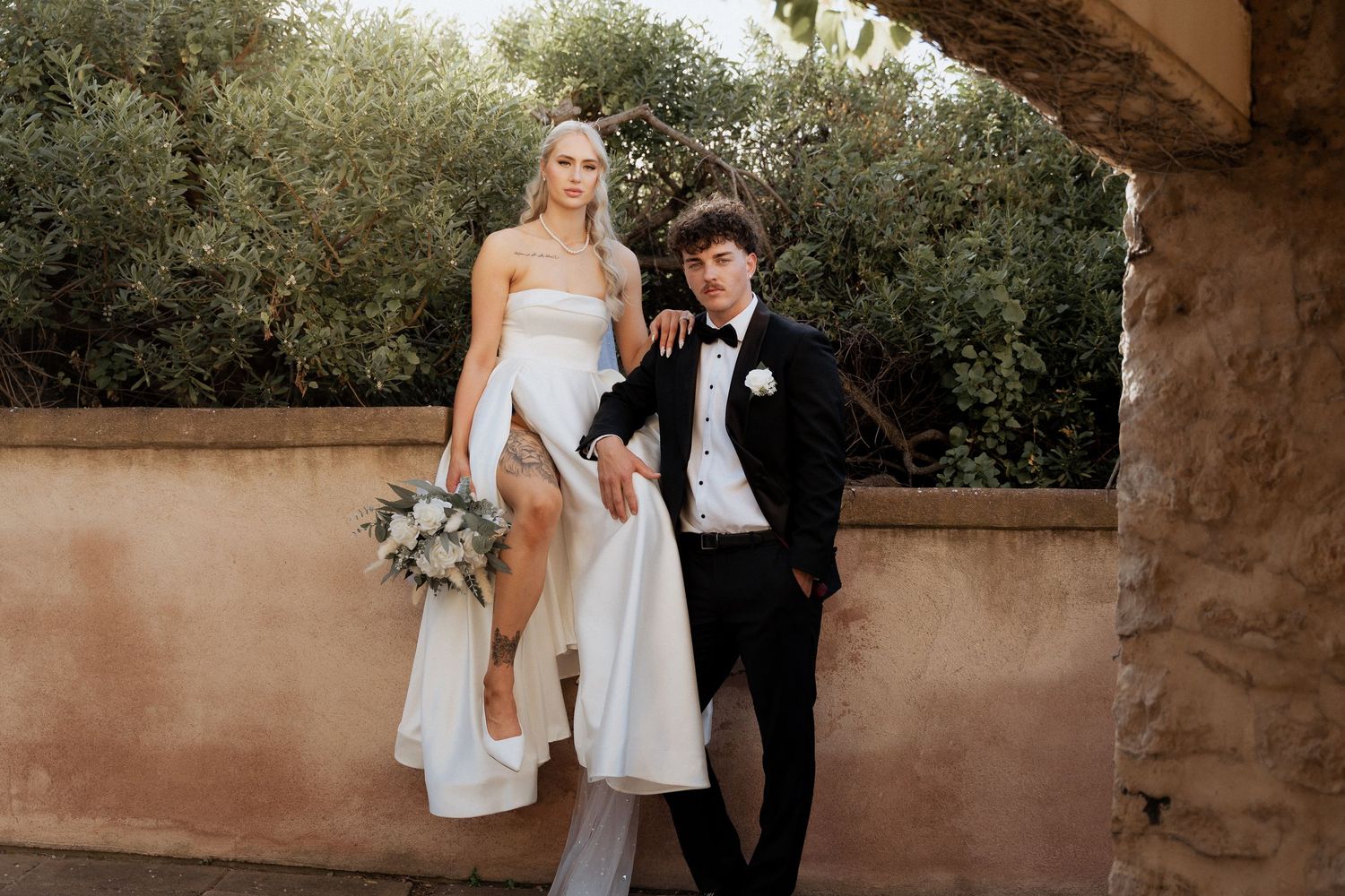A couple in formal wedding attire poses together in a sunlit garden setting with stone architecture.