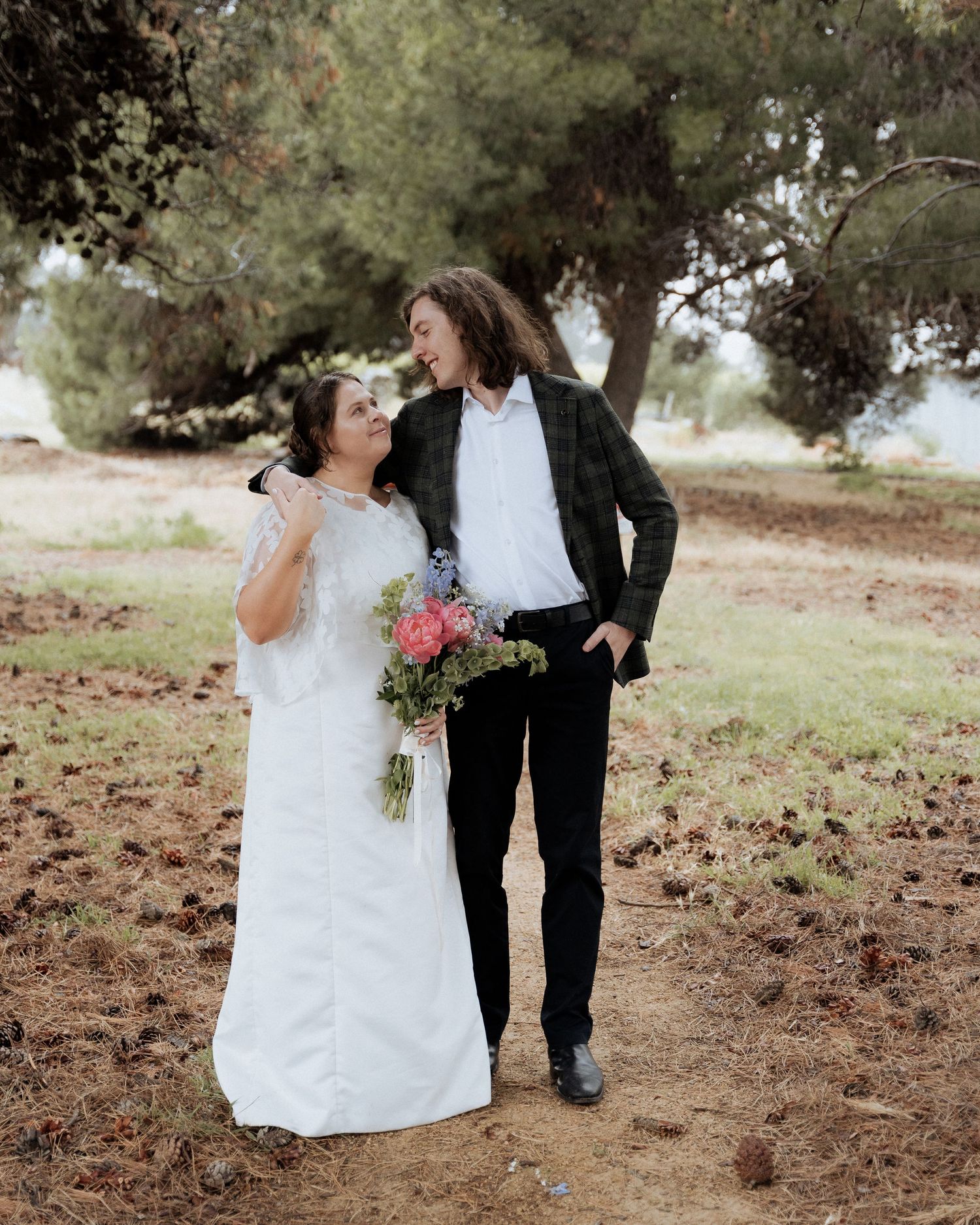 A smiling bride and groom walk together among trees, bride holding a colorful pink and blue bouquet at Ivy Brook Farm by Braytmedia