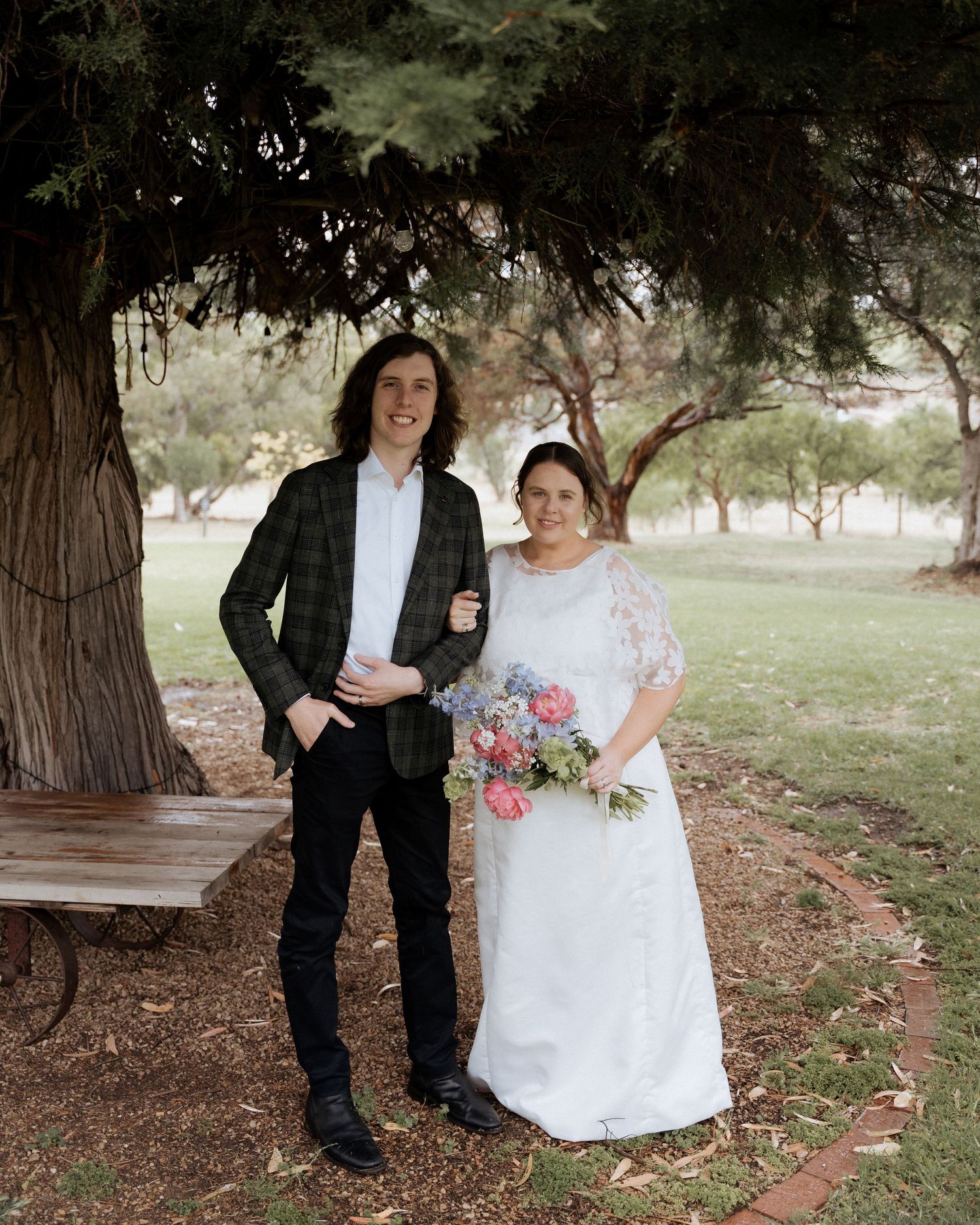 A bride and groom pose together under a large tree beside a rustic wooden bench outdoors at Ivy Brook Farm by Braytmedia