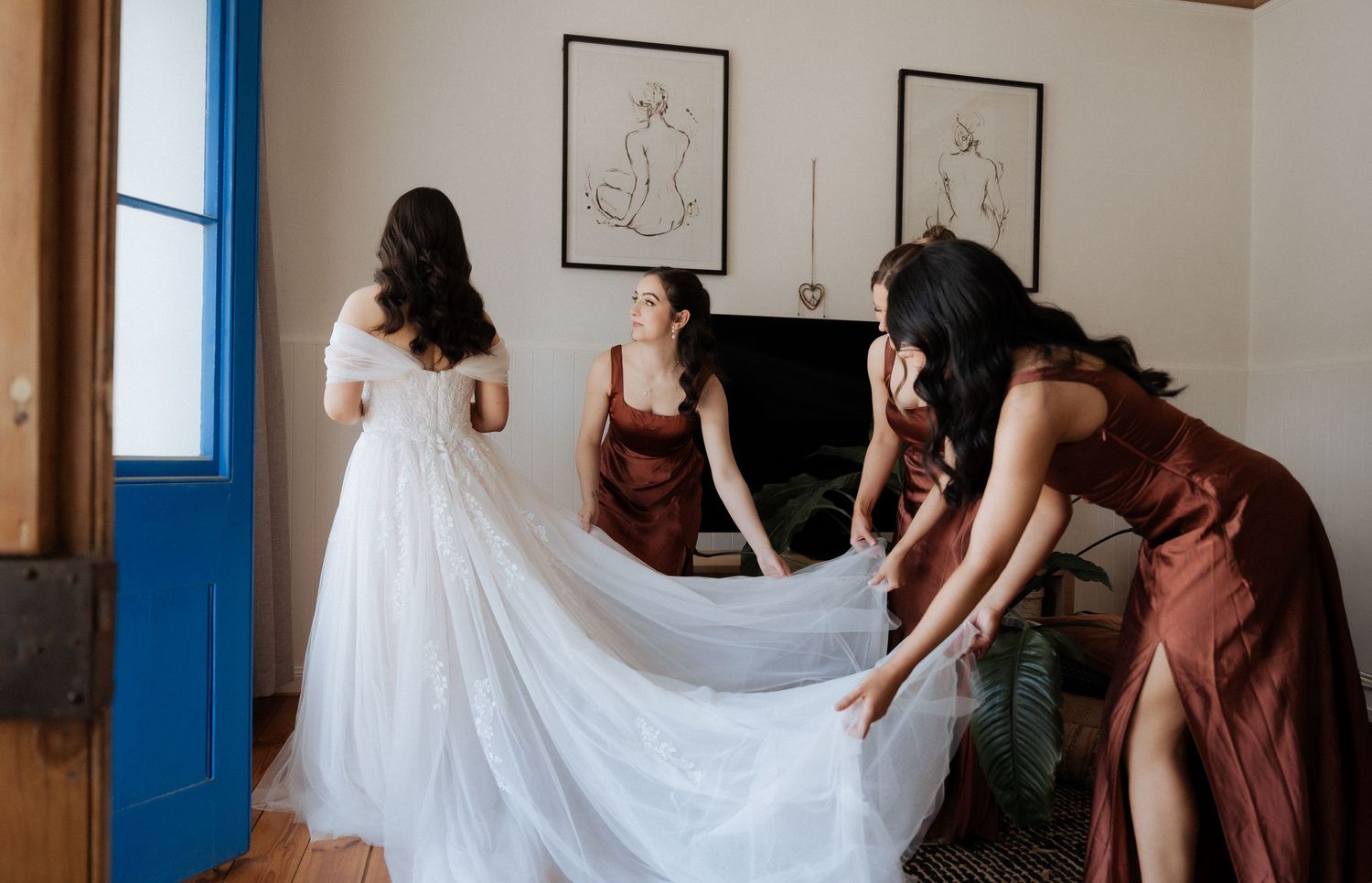 A wedding party helps adjust the bride's flowing white dress in a bright hallway.