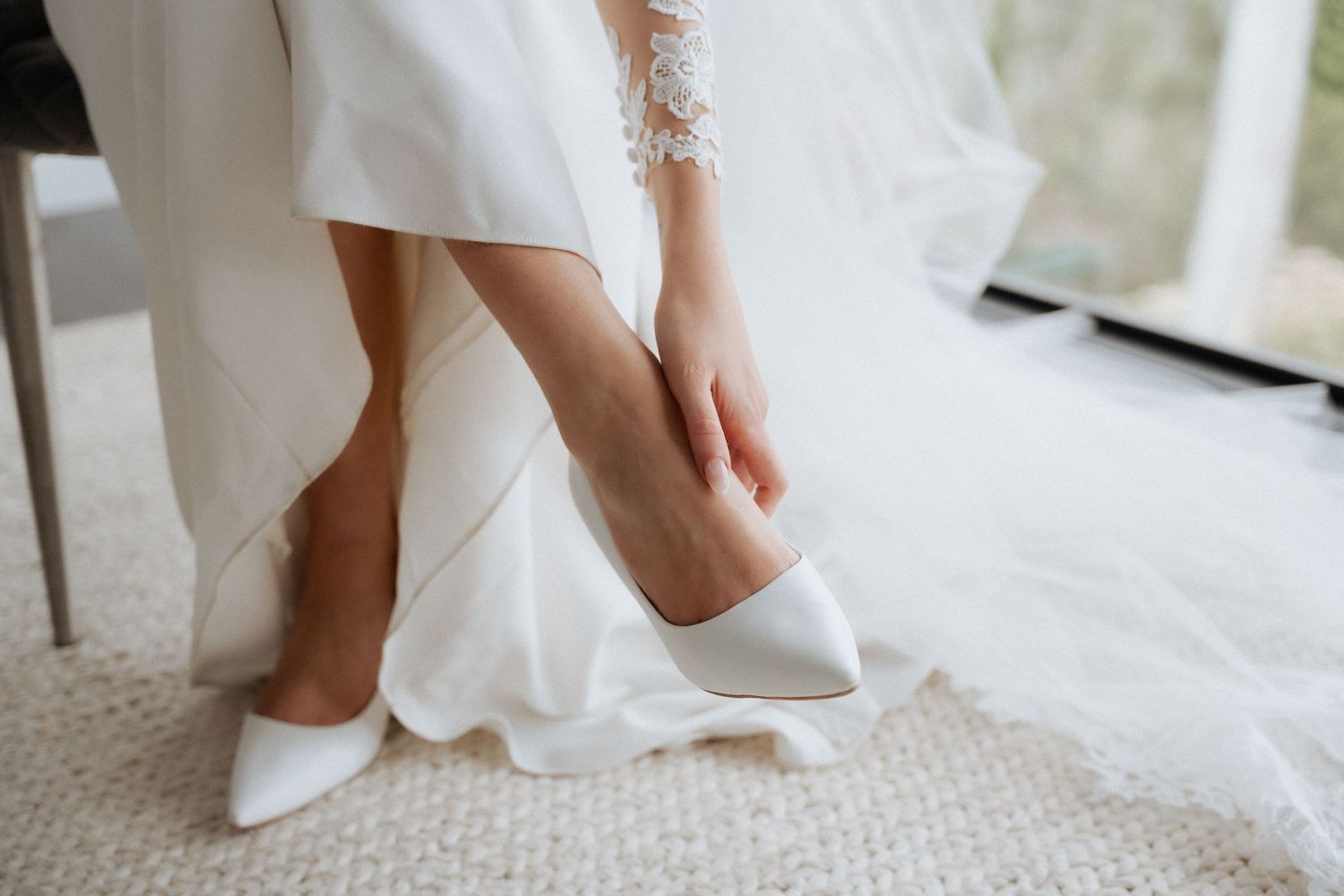 Close-up of bride putting on white satin pointed-toe heels while wearing a lace long-sleeve wedding gown by braytmedia adelaide wedding