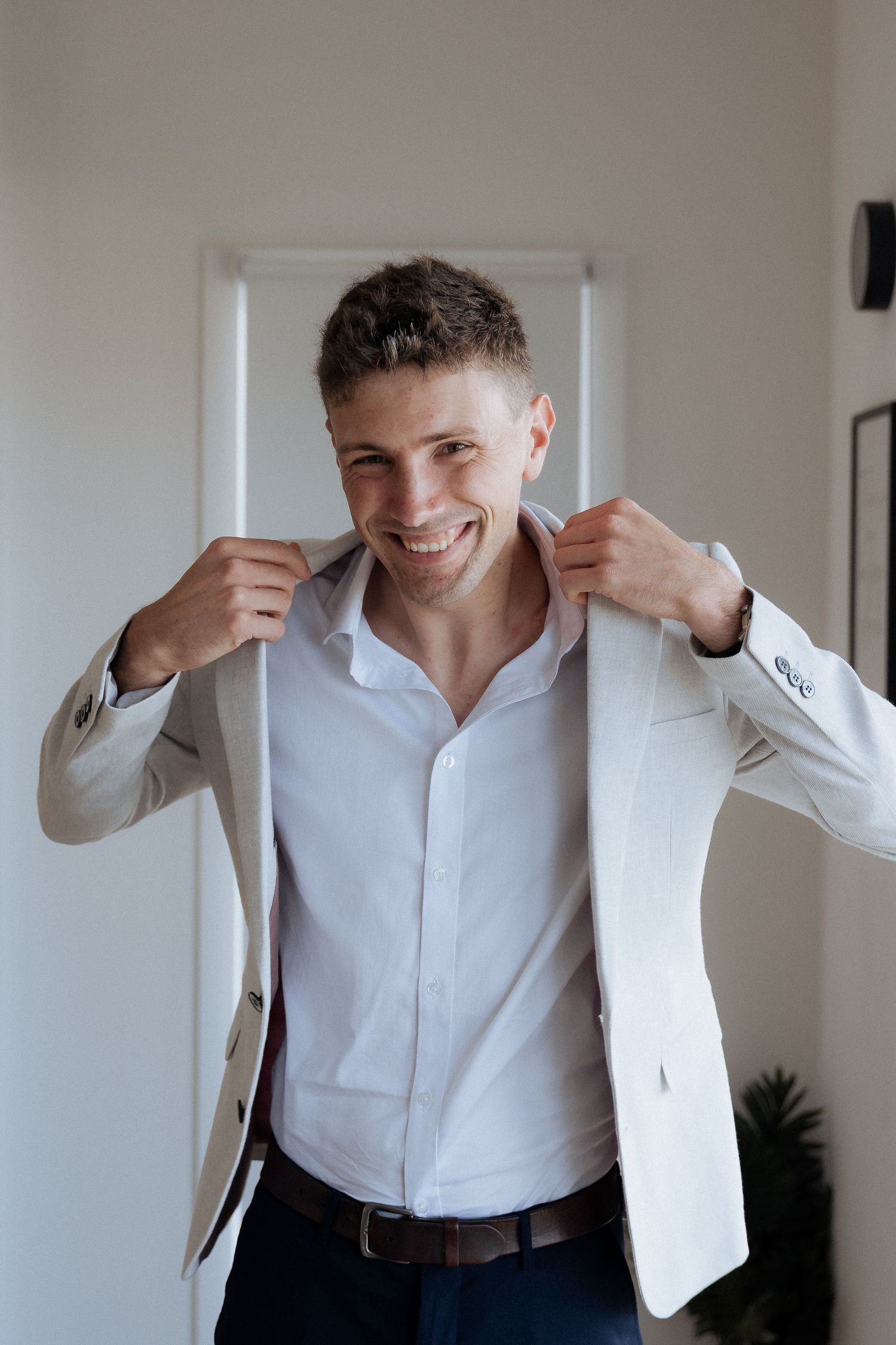 A person in a white blazer and dress shirt adjusts their collar while smiling.