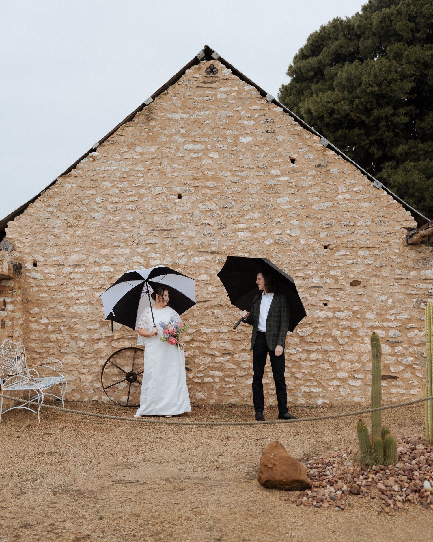 A bride and groom stand outside a rustic stone barn holding black umbrellas on a cloudy day at IvyBrook Farm Adelaide by braytmedia