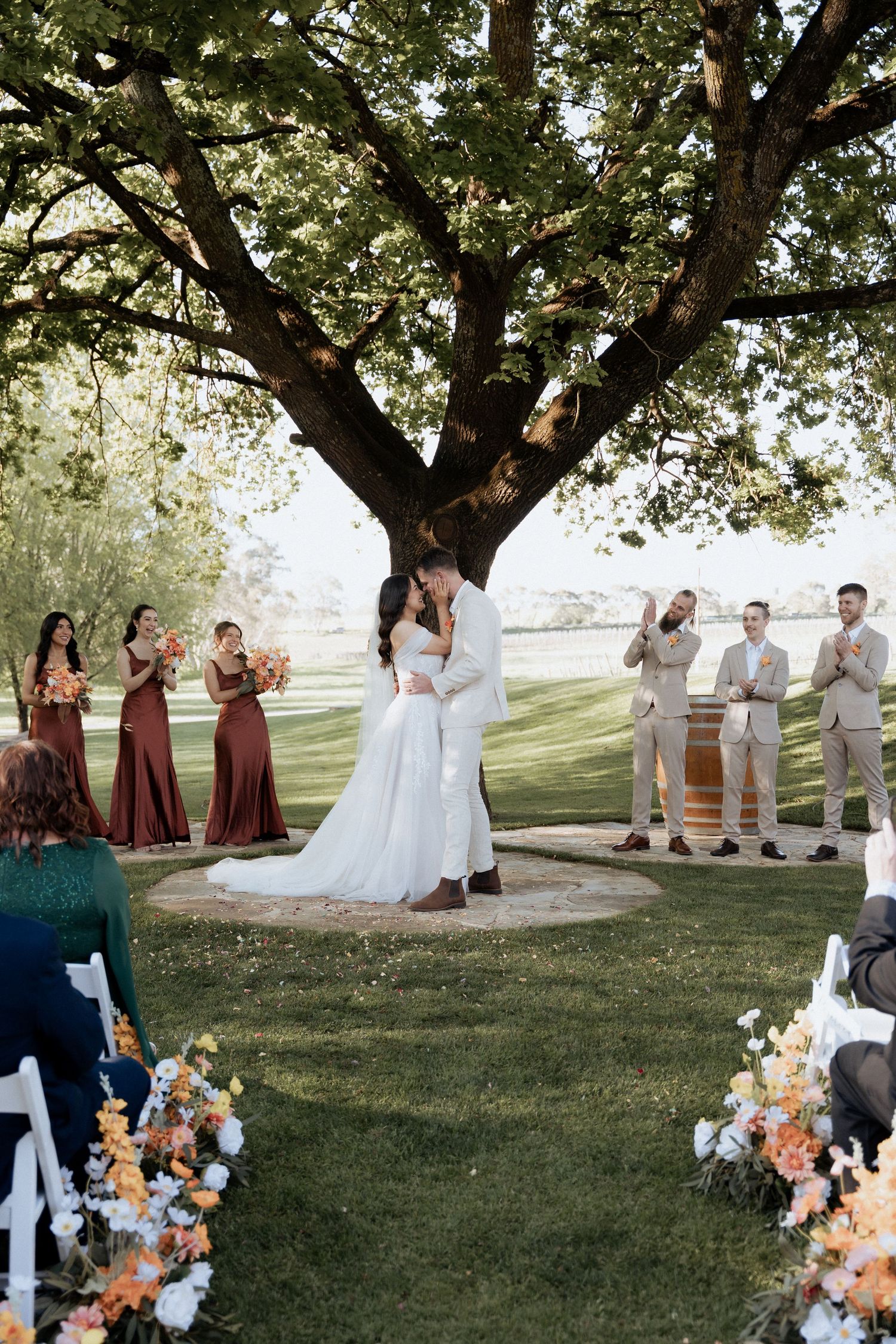 An outdoor wedding ceremony under a large tree with bridesmaids in burgundy dresses and groomsmen in beige suits.
