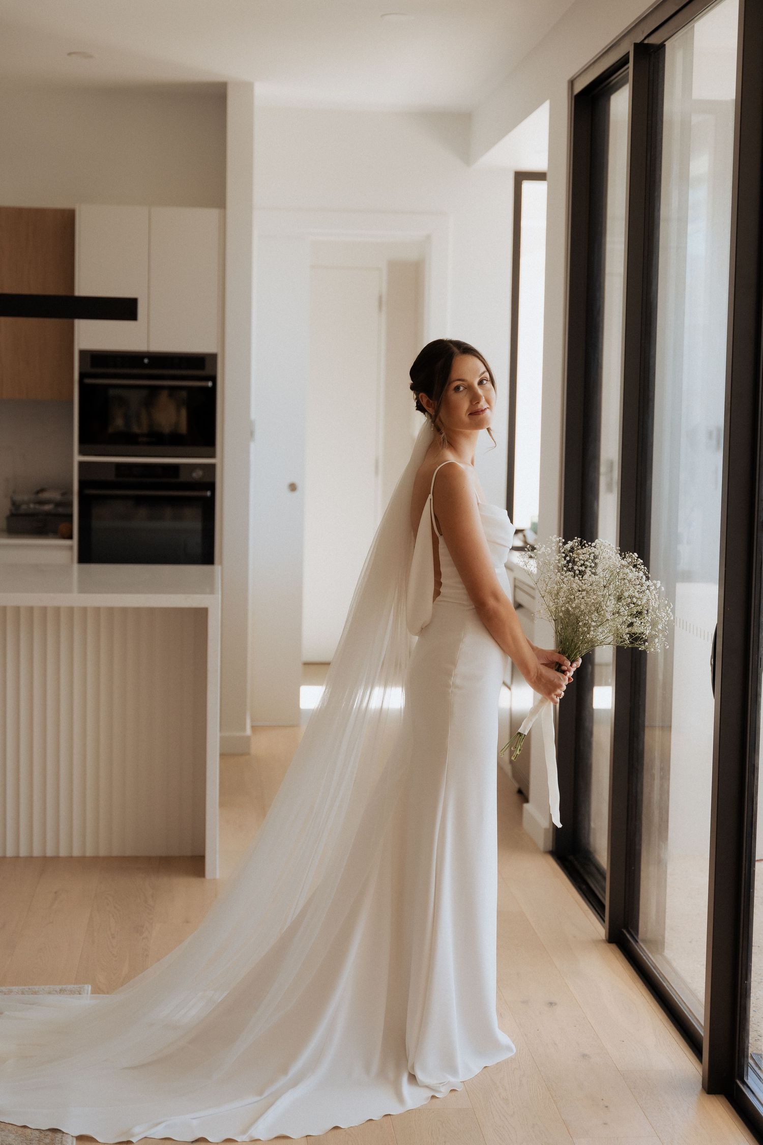 A graceful white wedding gown with a flowing train photographed against modern interior windows.