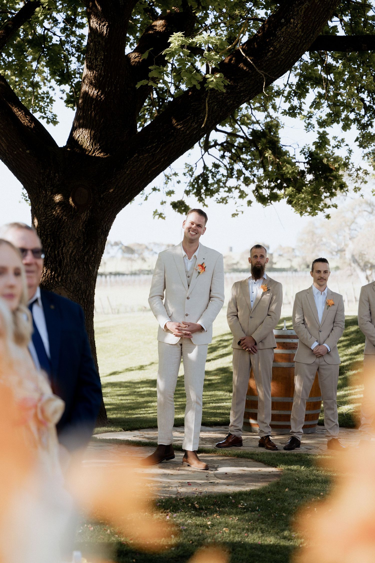 Groomsmen in light beige suits sit on wooden benches under a large tree during an outdoor wedding ceremony.