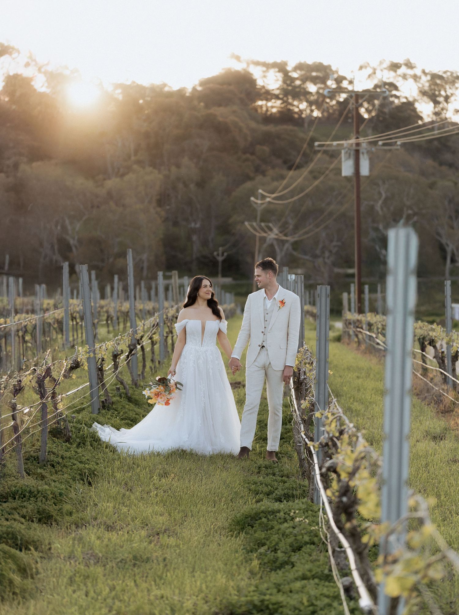 A couple walks through a vineyard at sunset with golden light filtering through the trees.