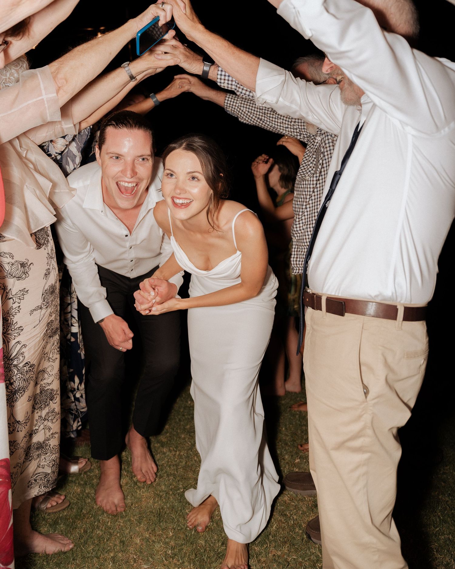 Wedding reception guests dancing together under string lights on grass in outdoor celebration.