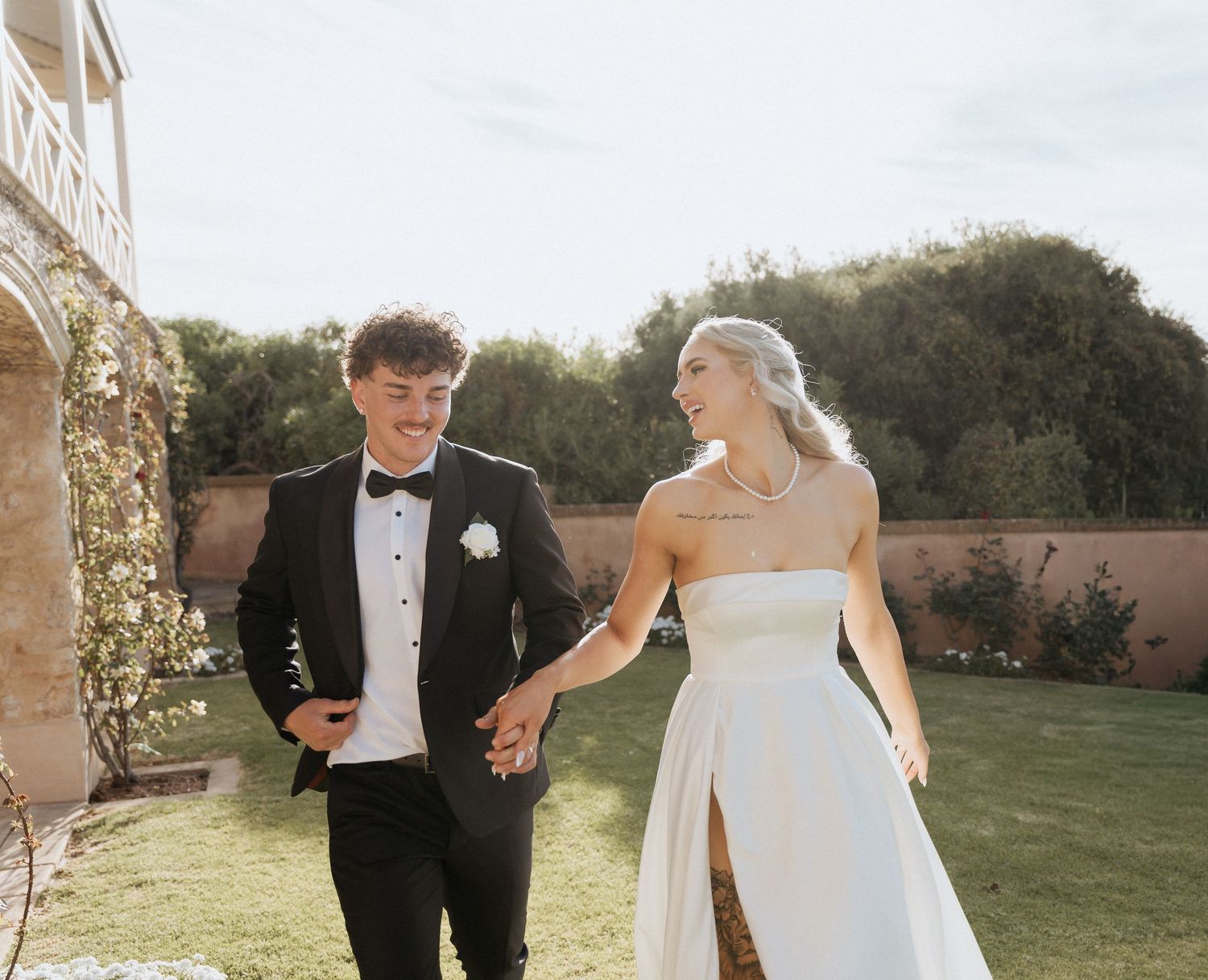 Newlyweds walk hand in hand through a lush garden pathway during golden hour.