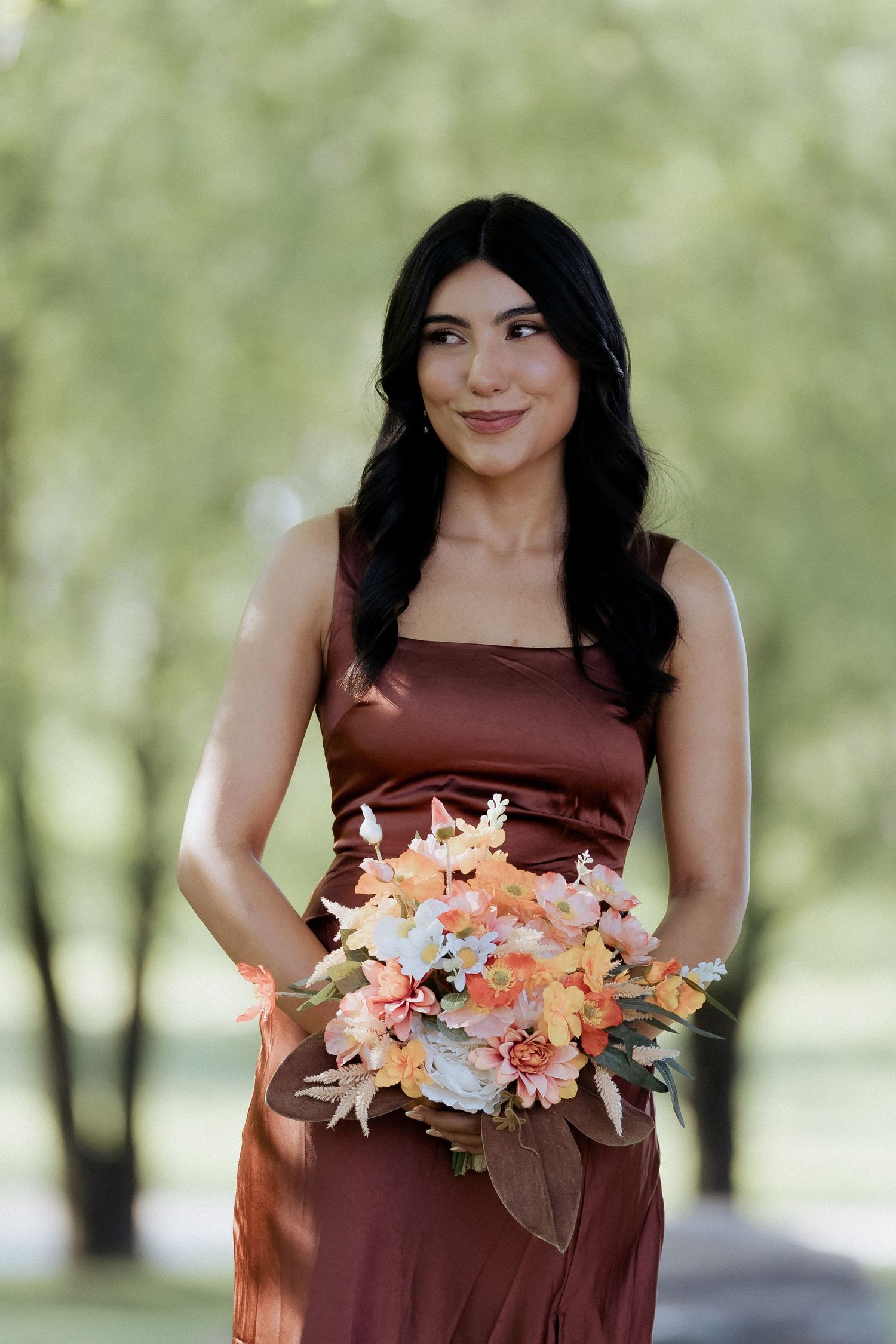 A person in a brown dress holds a colorful bouquet of peach and pink flowers against a blurred outdoor background.