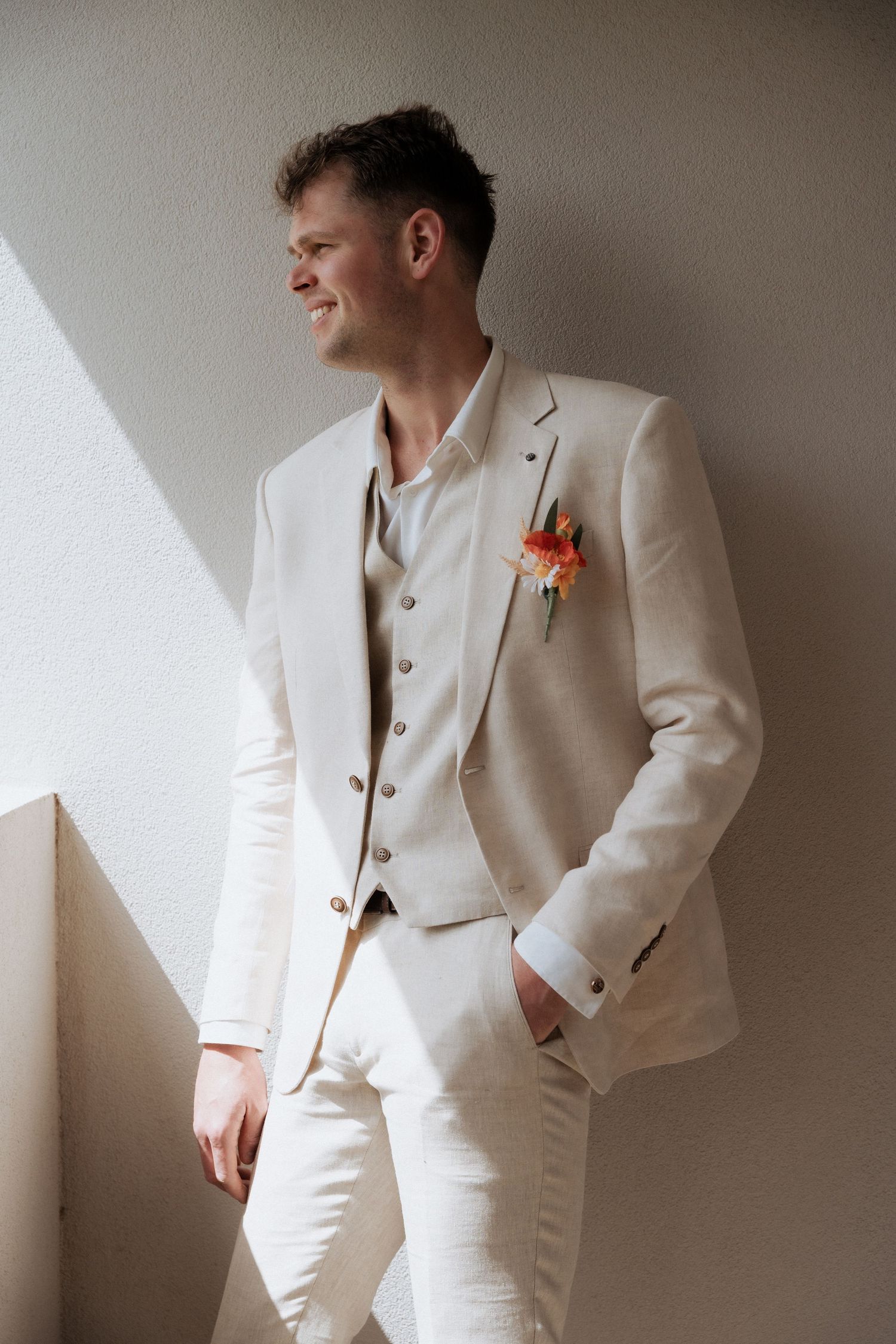 A person poses in a crisp white suit with orange boutonniere against a neutral wall in a series of fashion portraits.