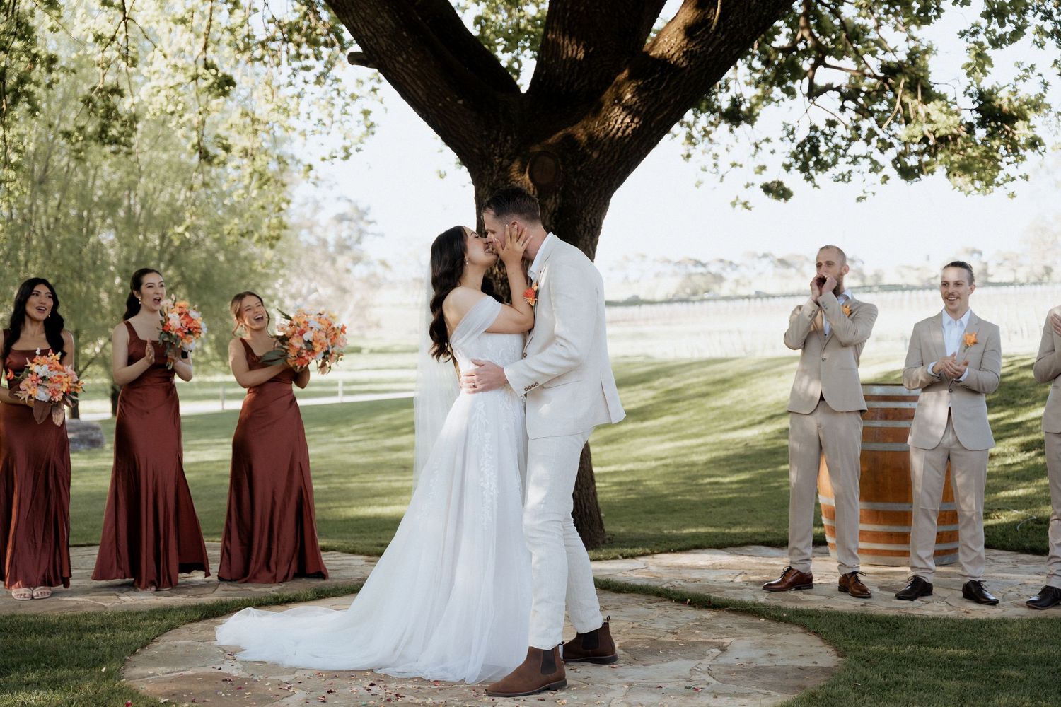 Wedding celebration with guests gathered under a large tree during an outdoor ceremony with burgundy and white attire.