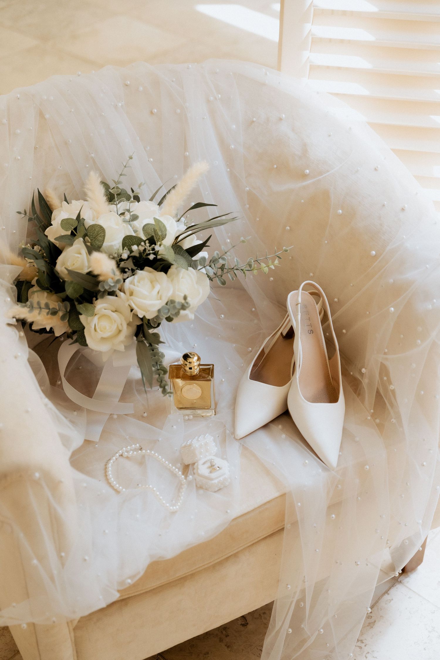 White wedding shoes and floral bouquet arranged with delicate tulle veil on light wooden surface.
