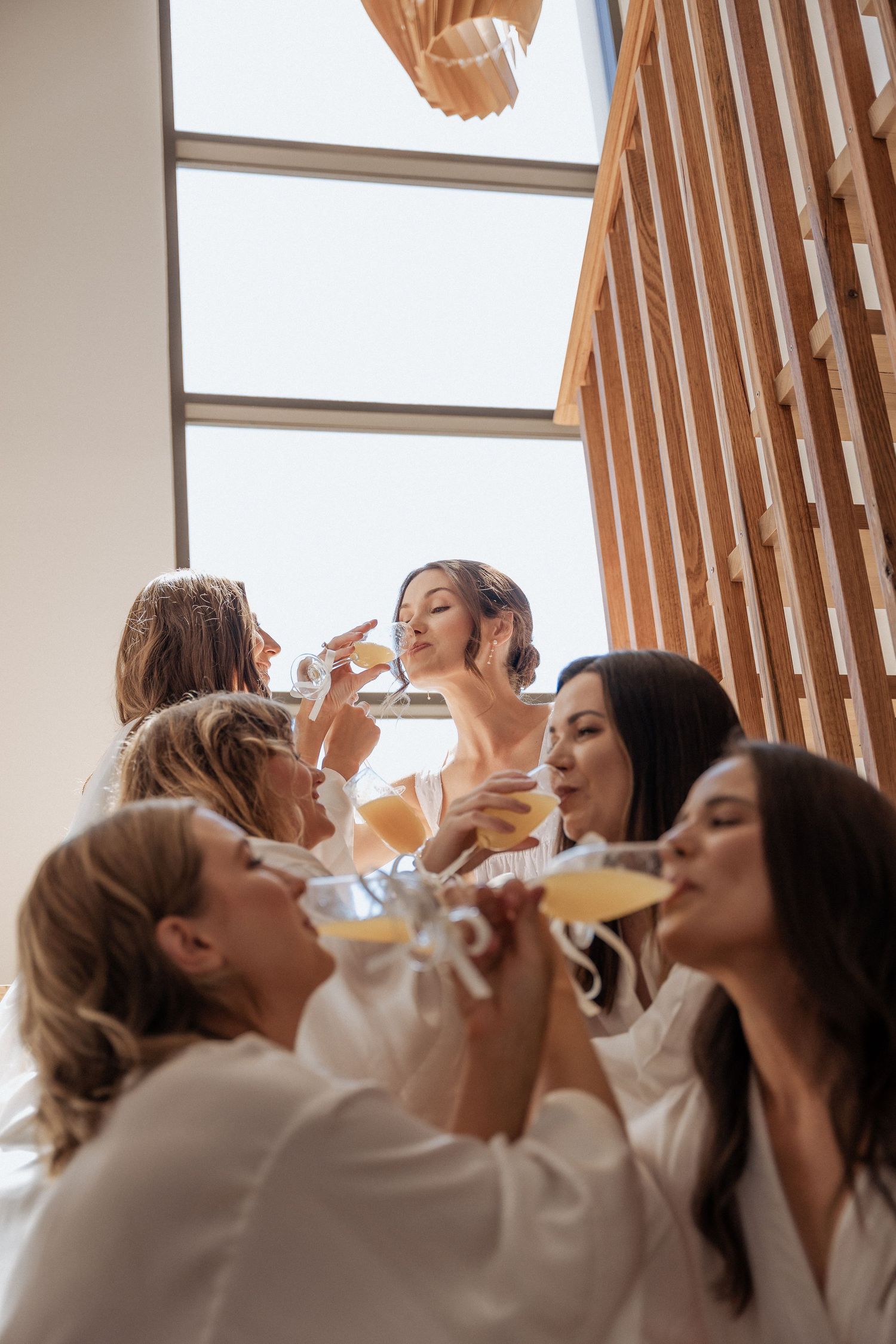 Group of bridesmaids in white robes toasting with champagne glasses by a window.