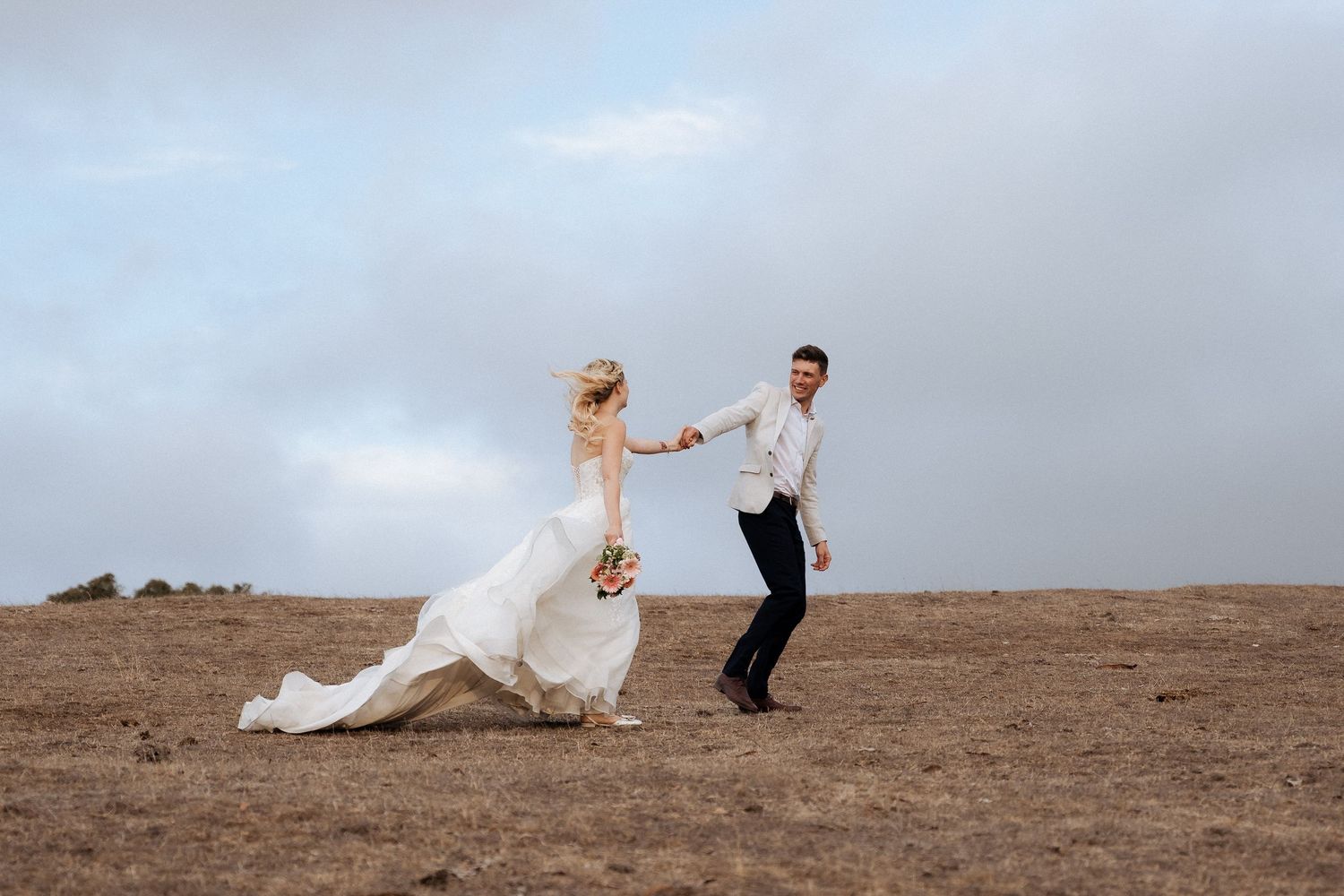 Wedding couple embracing on a scenic hilltop with dress flowing in the wind. Captured by braytmedia