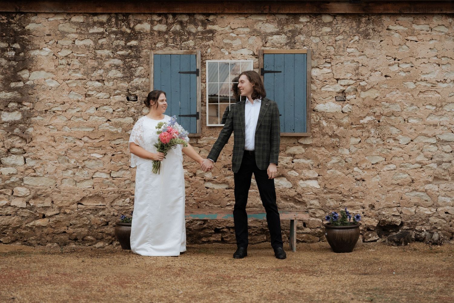 A bride holding a pink bouquet and groom hold hands outside a rustic stone building with blue shutters Ivybrook Farm images by braytmedia
