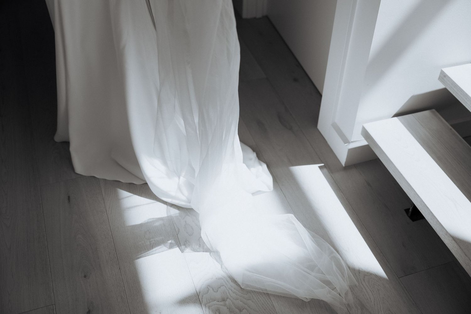 A flowing white wedding dress train spreads across a wooden floor in dramatic natural light.