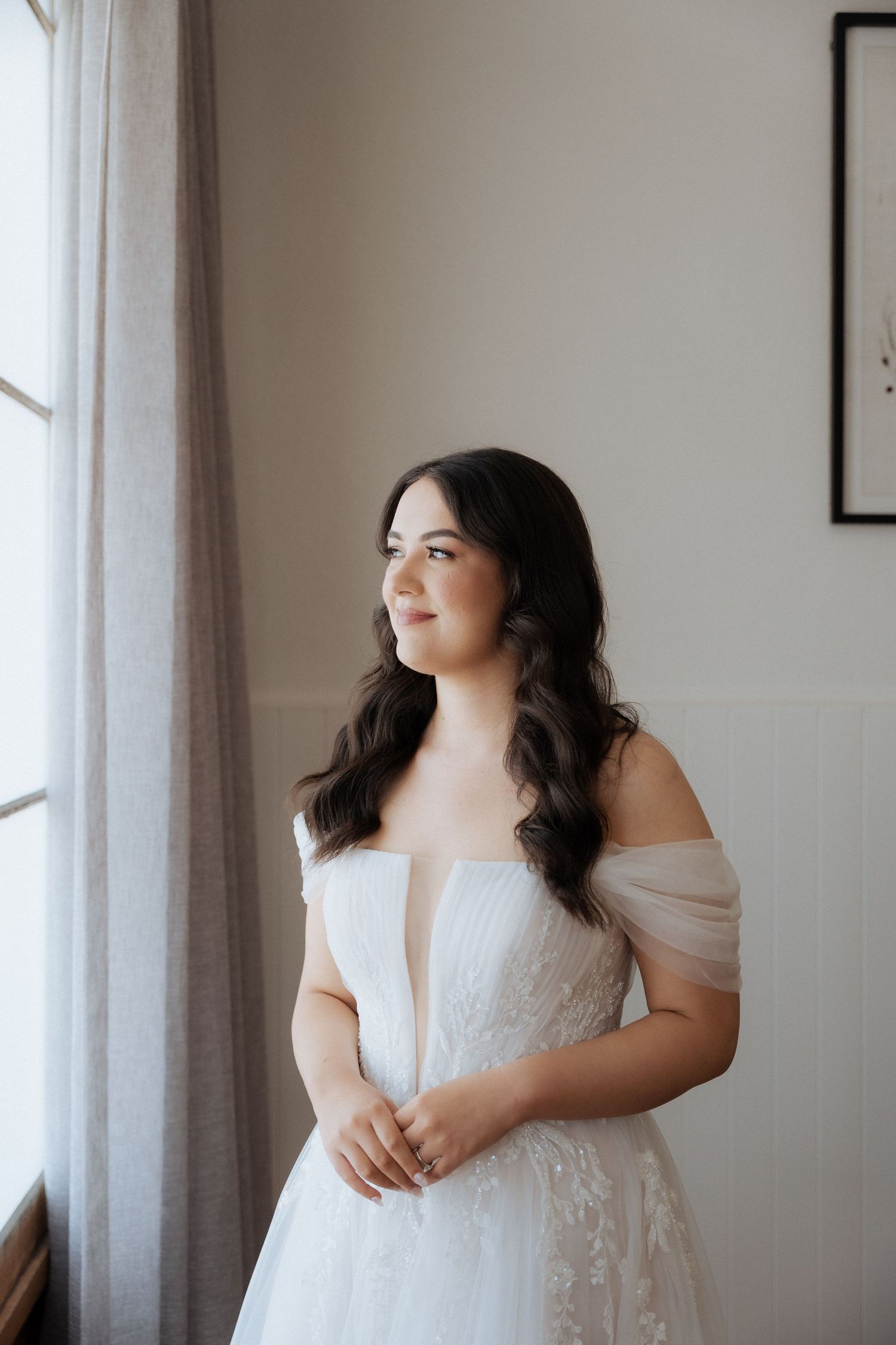 A bride in an off-shoulder white lace wedding gown gazes out a window in soft natural light.