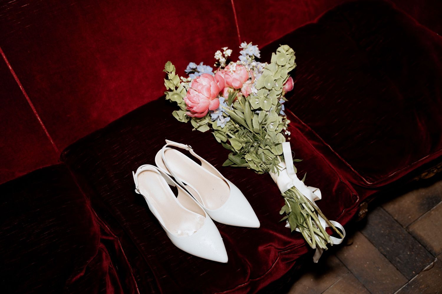 White slingback bridal heels and a pink and blue floral bouquet rest on red velvet. by Braytmedia at Ivy Brook Farm