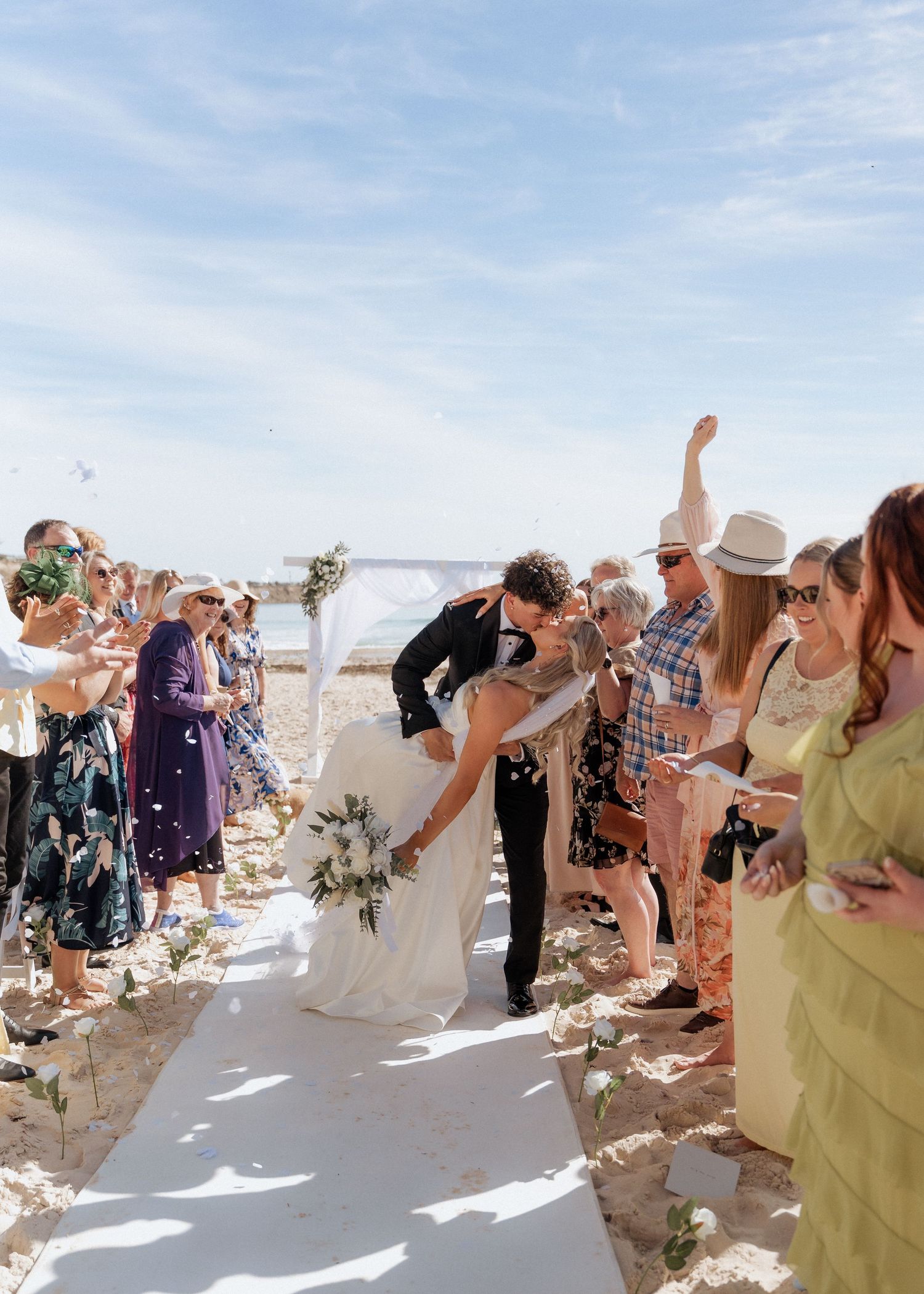 Wedding guests celebrate with raised arms on a sunny beach during an outdoor ceremony wedding on Pt willunga beach, images by braytmedia