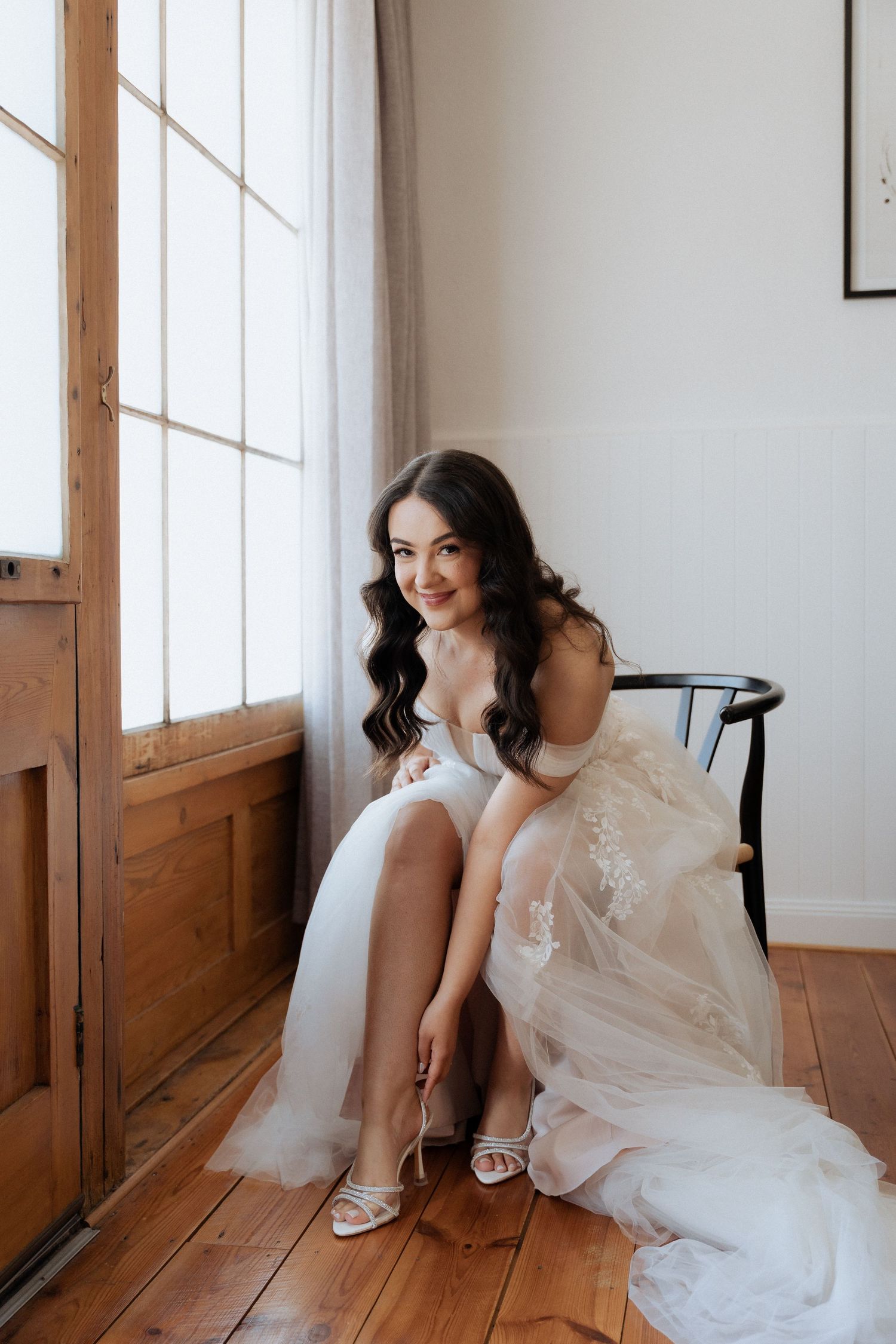 A bride in a flowing white dress sits gracefully on a wooden floor near large windows in a bright indoor setting.