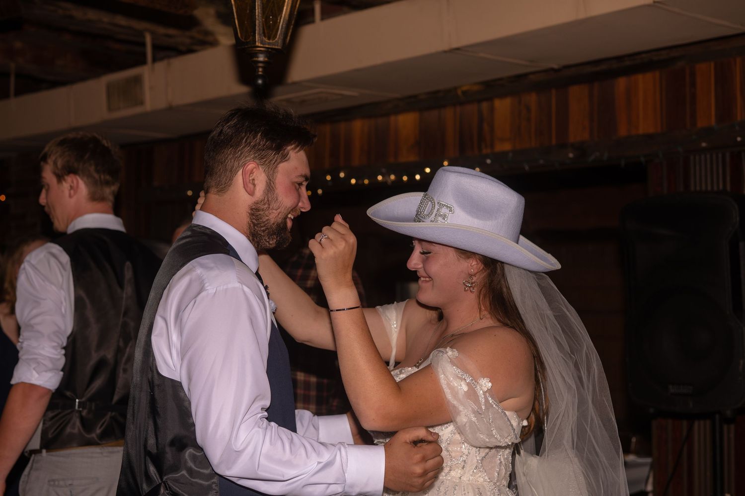 Bride in white dress and cowboy hat dances with groom in vest during rustic barn wedding reception.