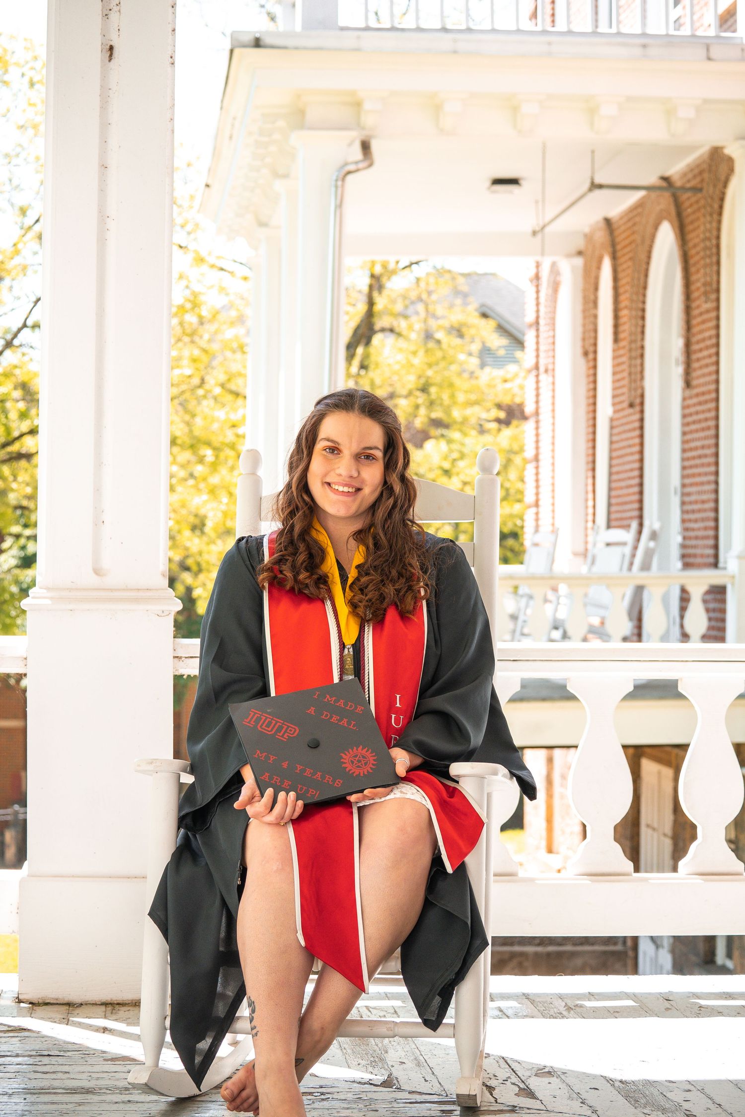 Graduate in black robe and red stole sits near white columns outdoors.