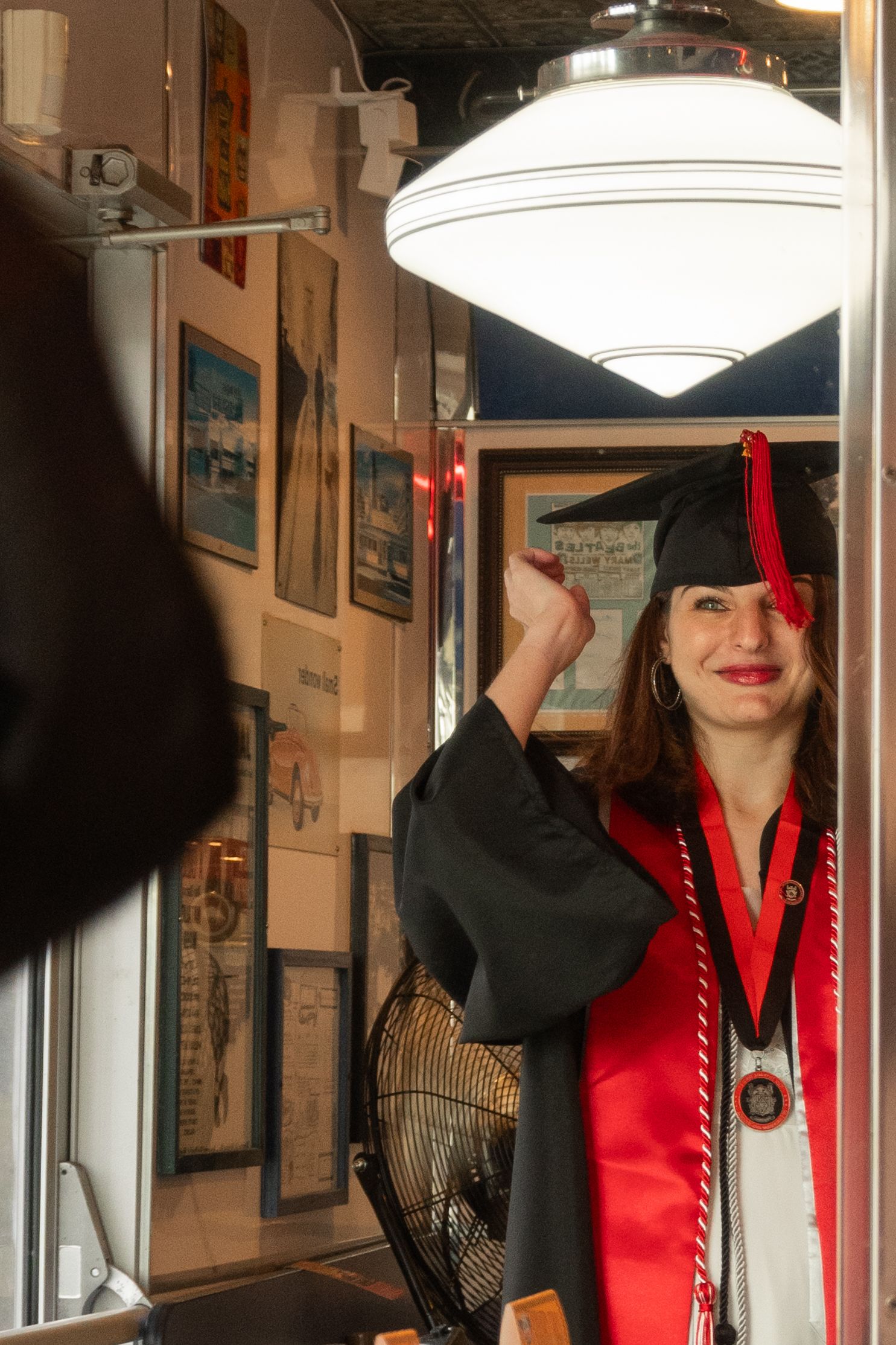 Graduate in black gown and red stole poses with graduation cap in checkerboard room.