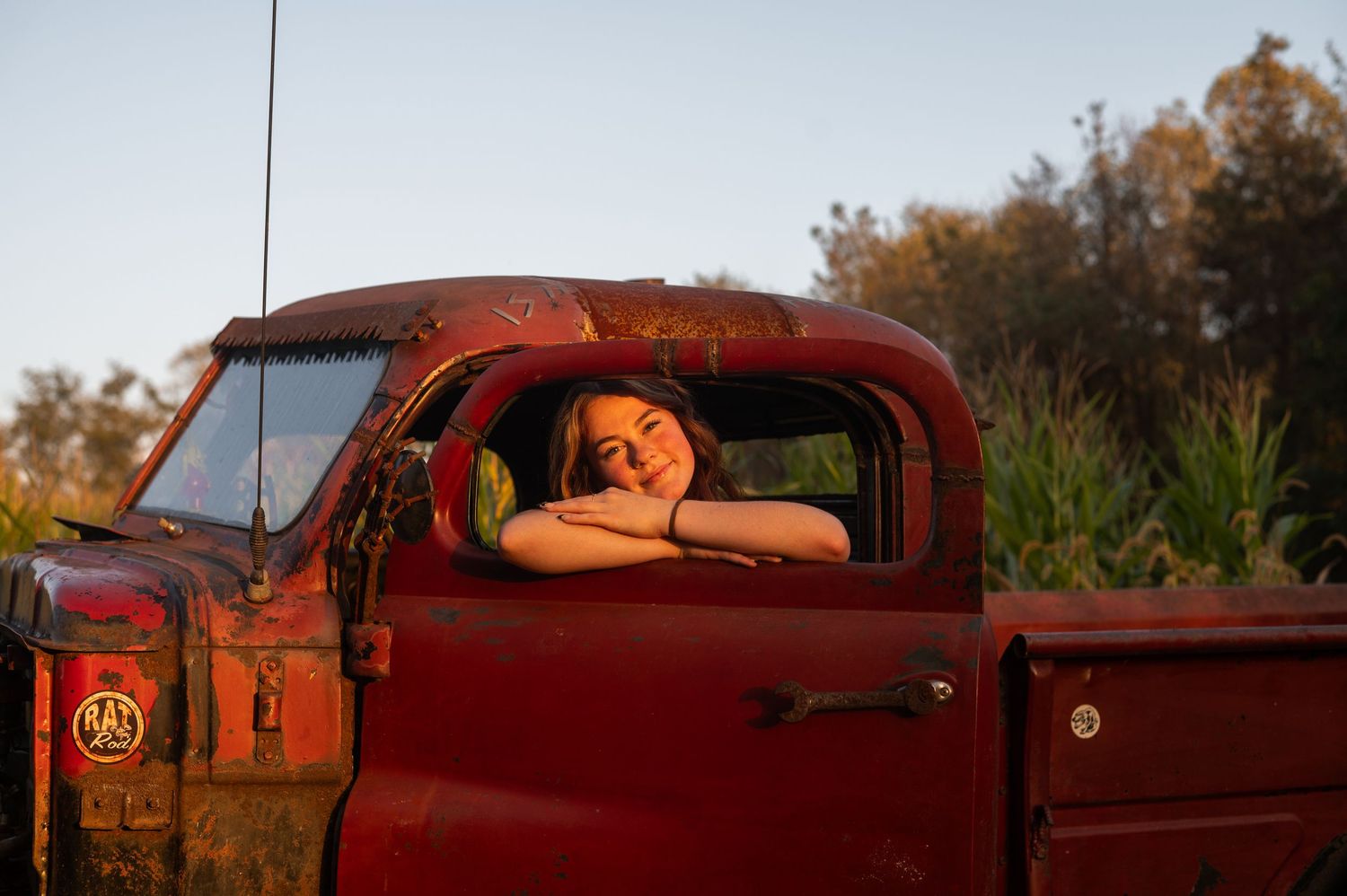 A vintage red pickup truck sits in golden sunlight with someone resting in the driver's window during sunset.