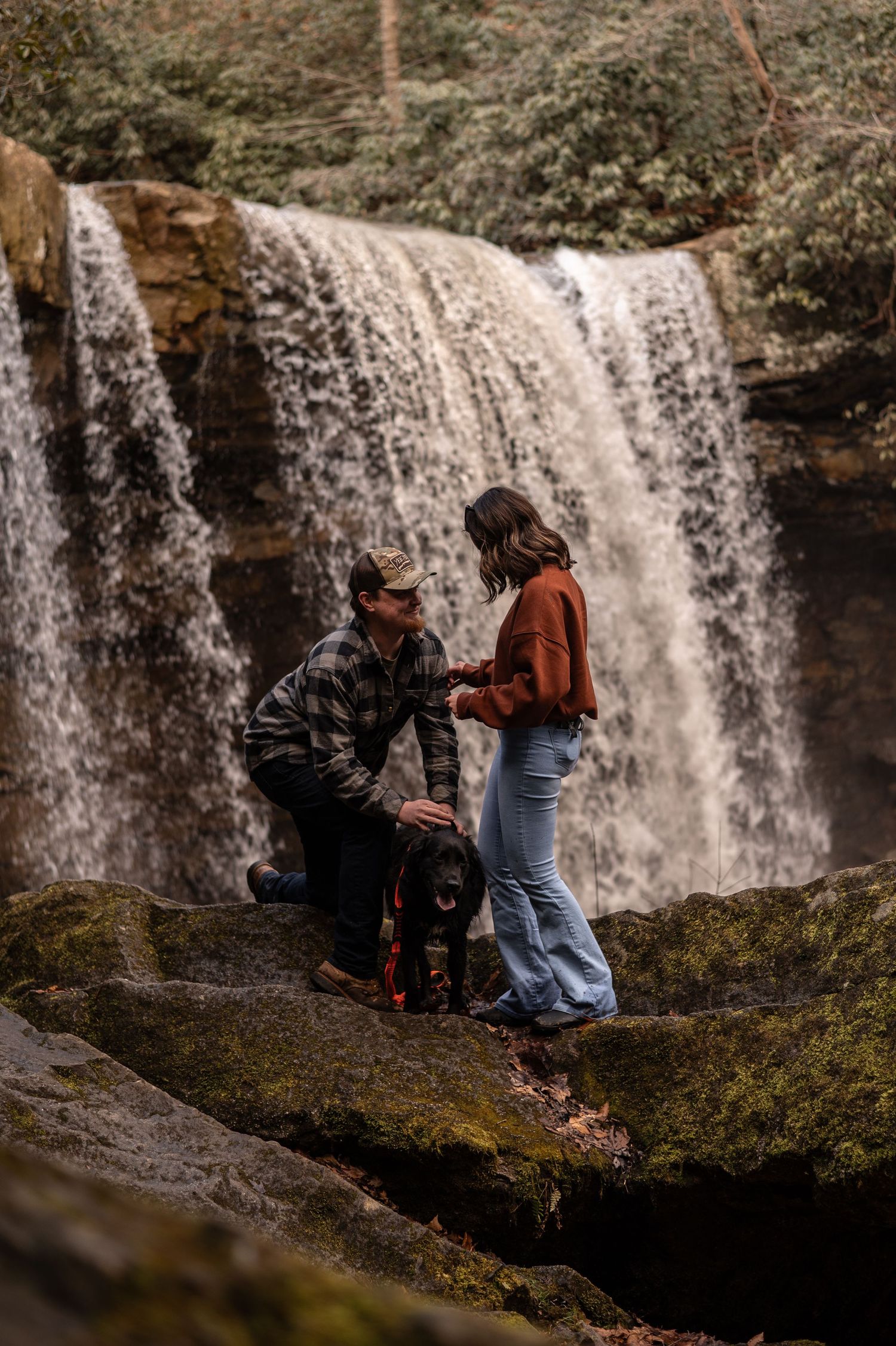 People sit on mossy rocks near a scenic waterfall in a peaceful natural setting.