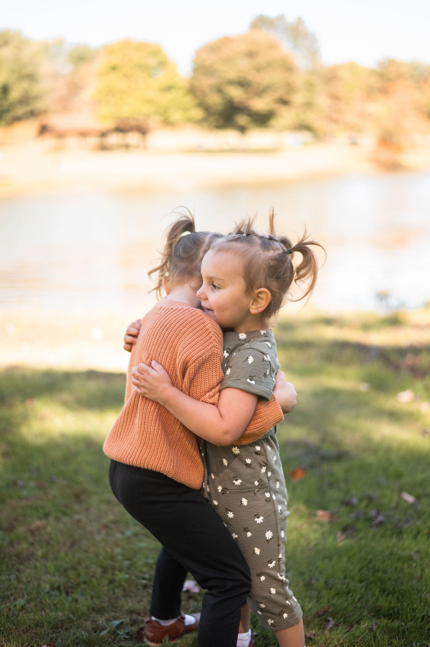 Two young toddler girls hugging outdoors near a lake, wearing casual clothes, with trees in the background.