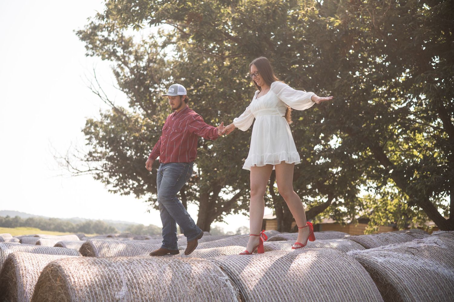 Two people hold hands while balancing on hay bales in a sunlit field during golden hour.