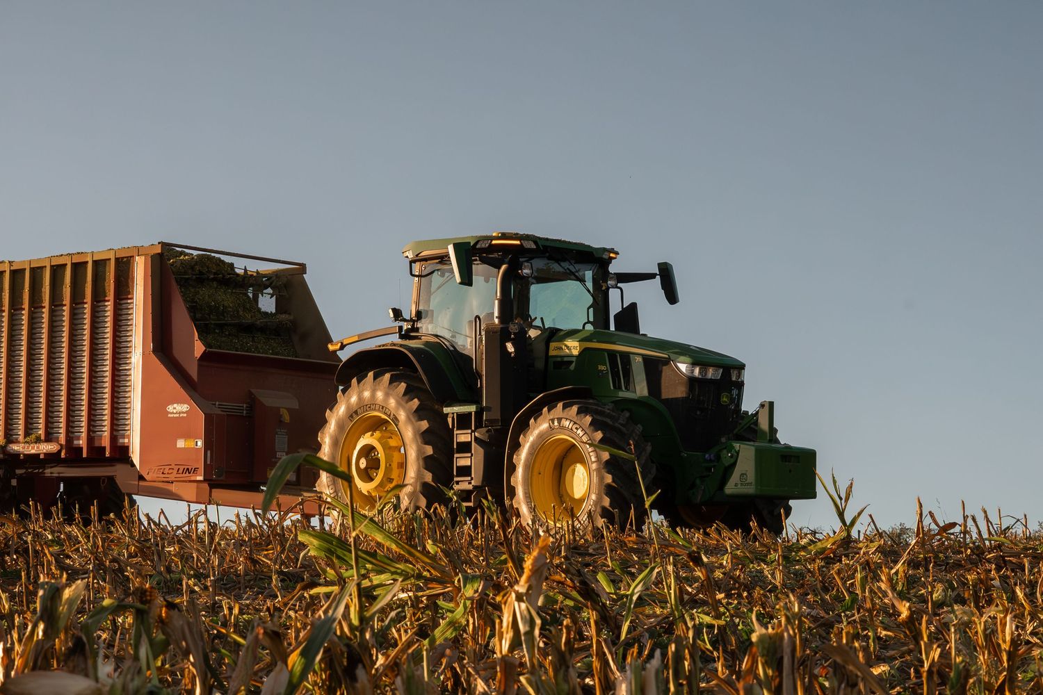 Silhouettes of agricultural machinery and trucks against a dusky sky in a harvested field.