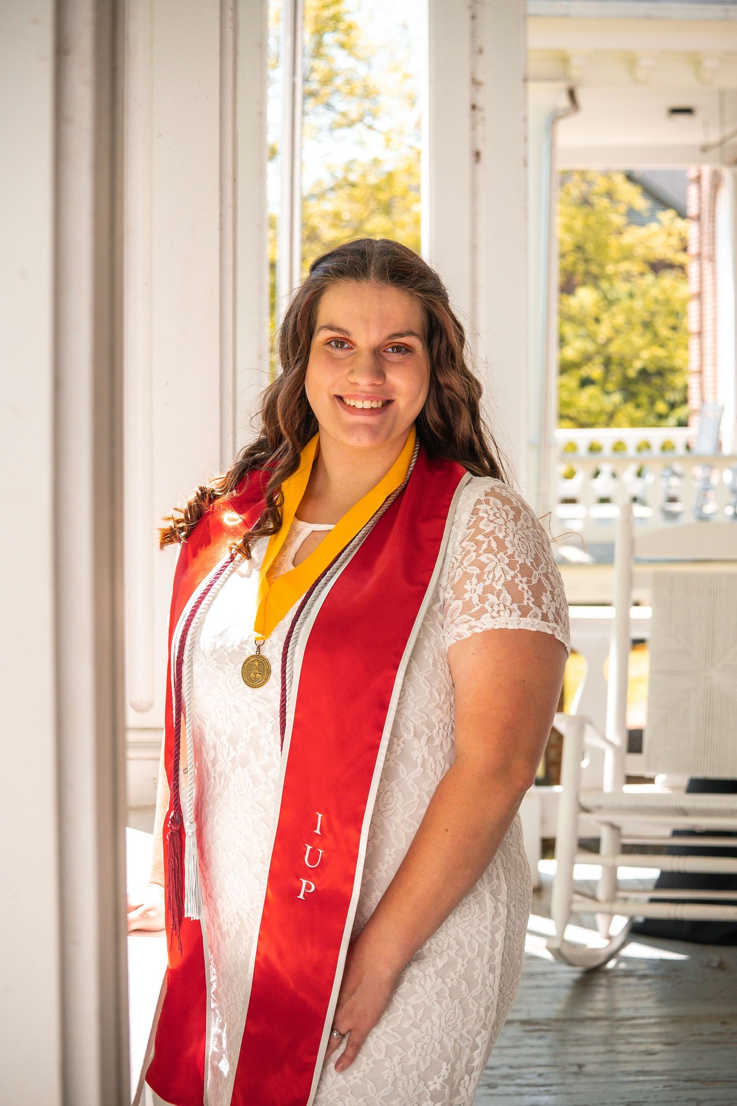 Graduate wearing white lace and red stole poses on a sunny porch.