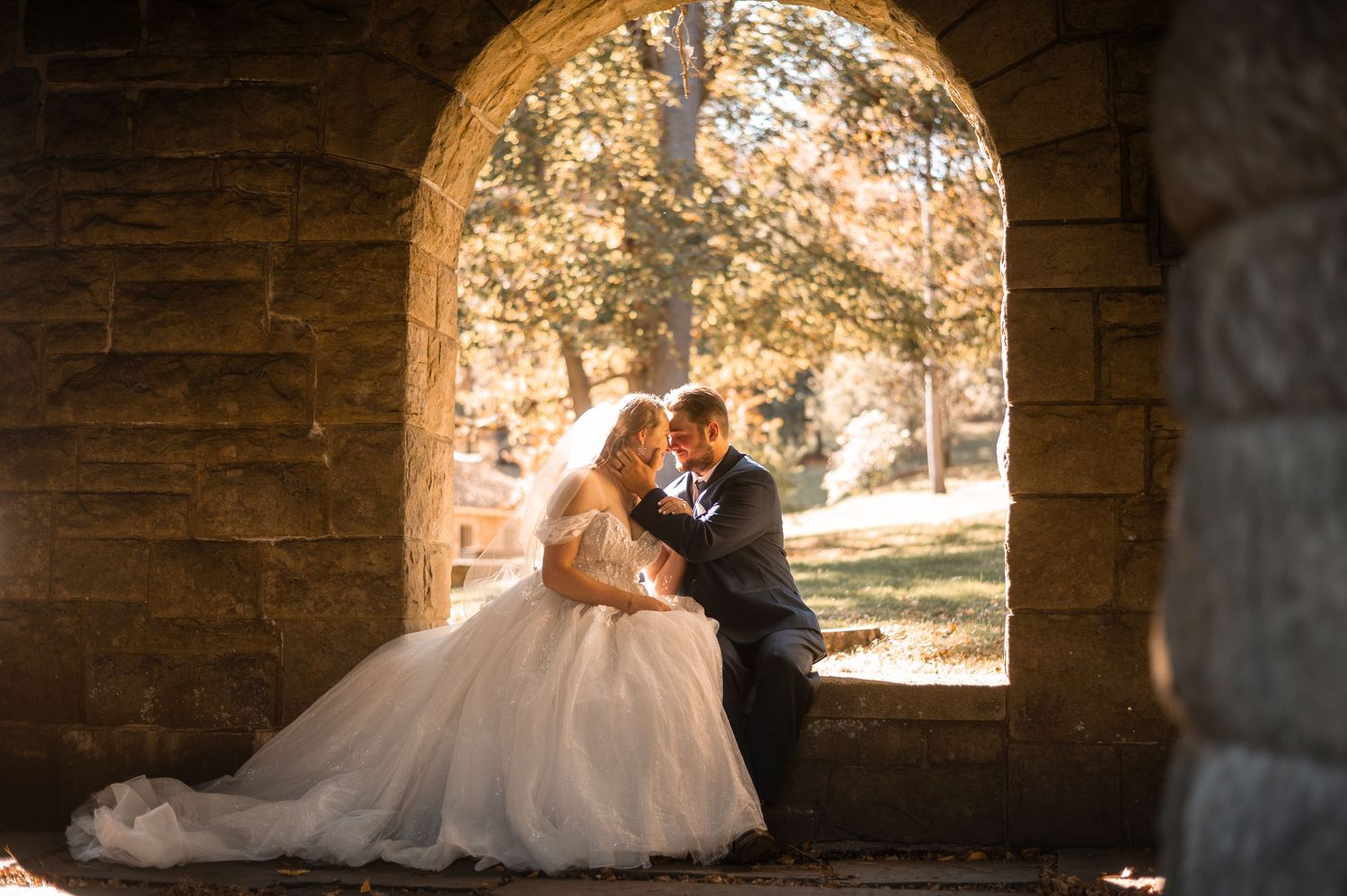 Romantic wedding couple embraces in stone archway with glowing autumn foliage in background.