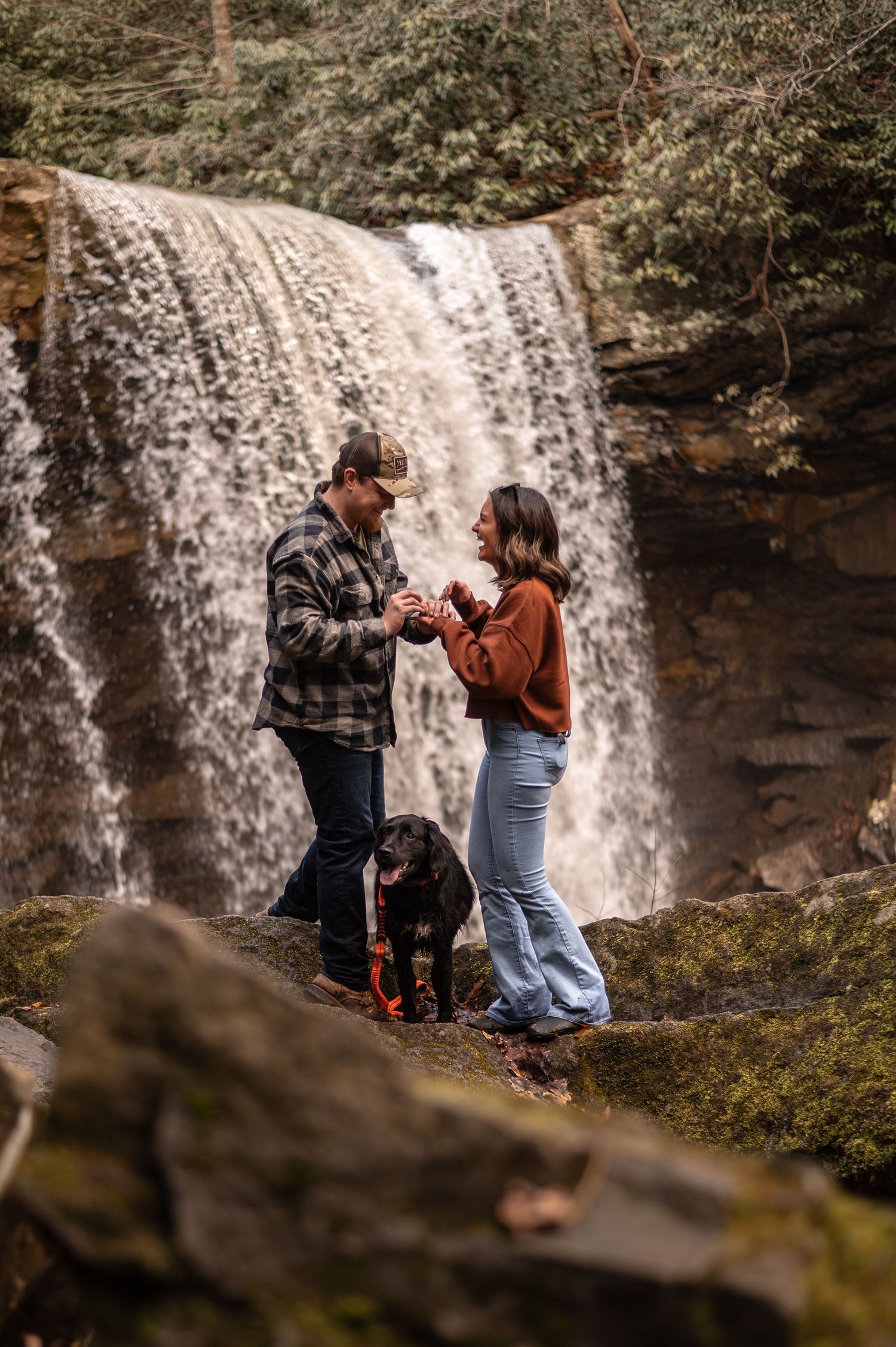 Two people share an intimate moment in front of a cascading waterfall in a scenic forest setting.