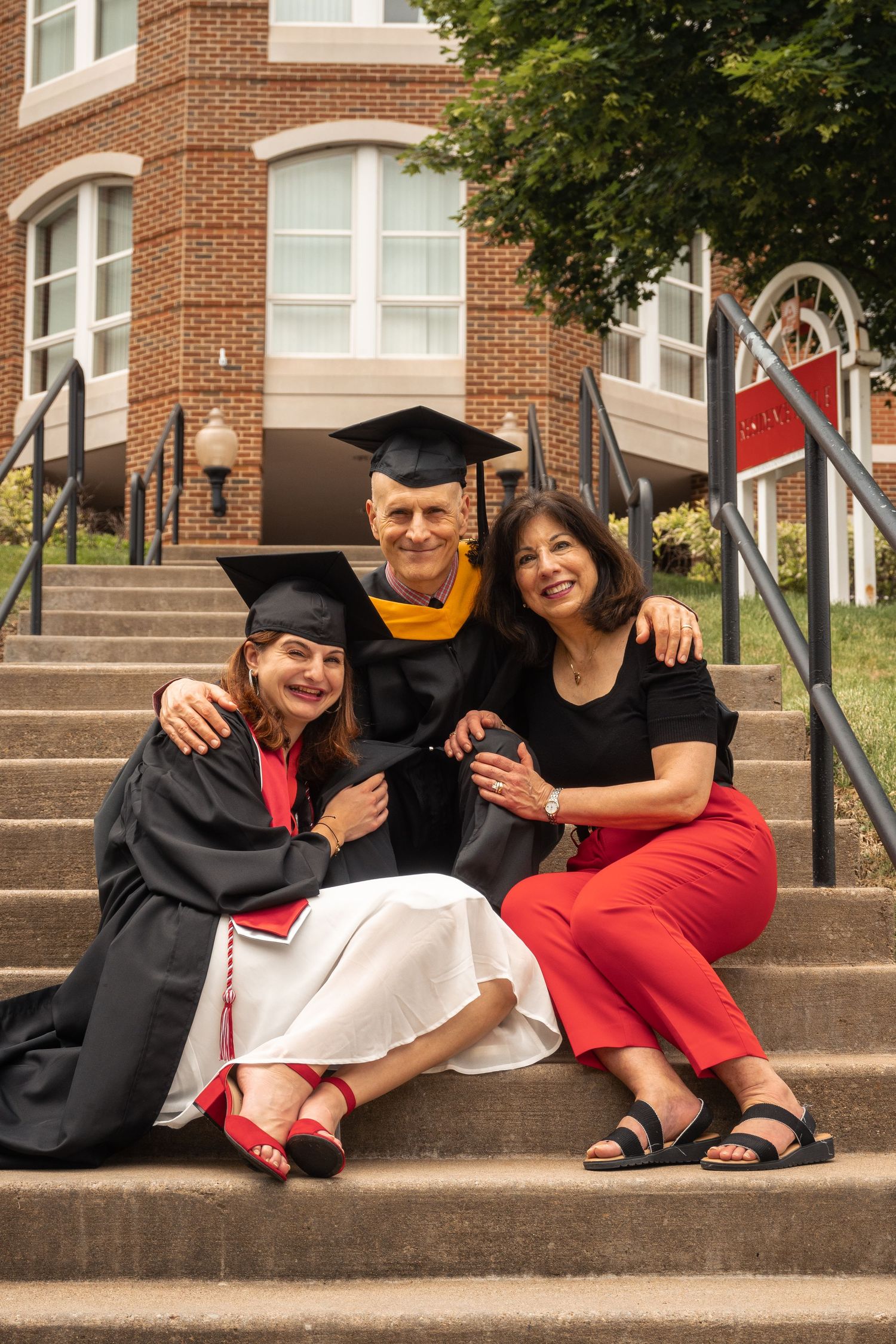 Graduates in black robes and colorful attire sit together on brick stairs.