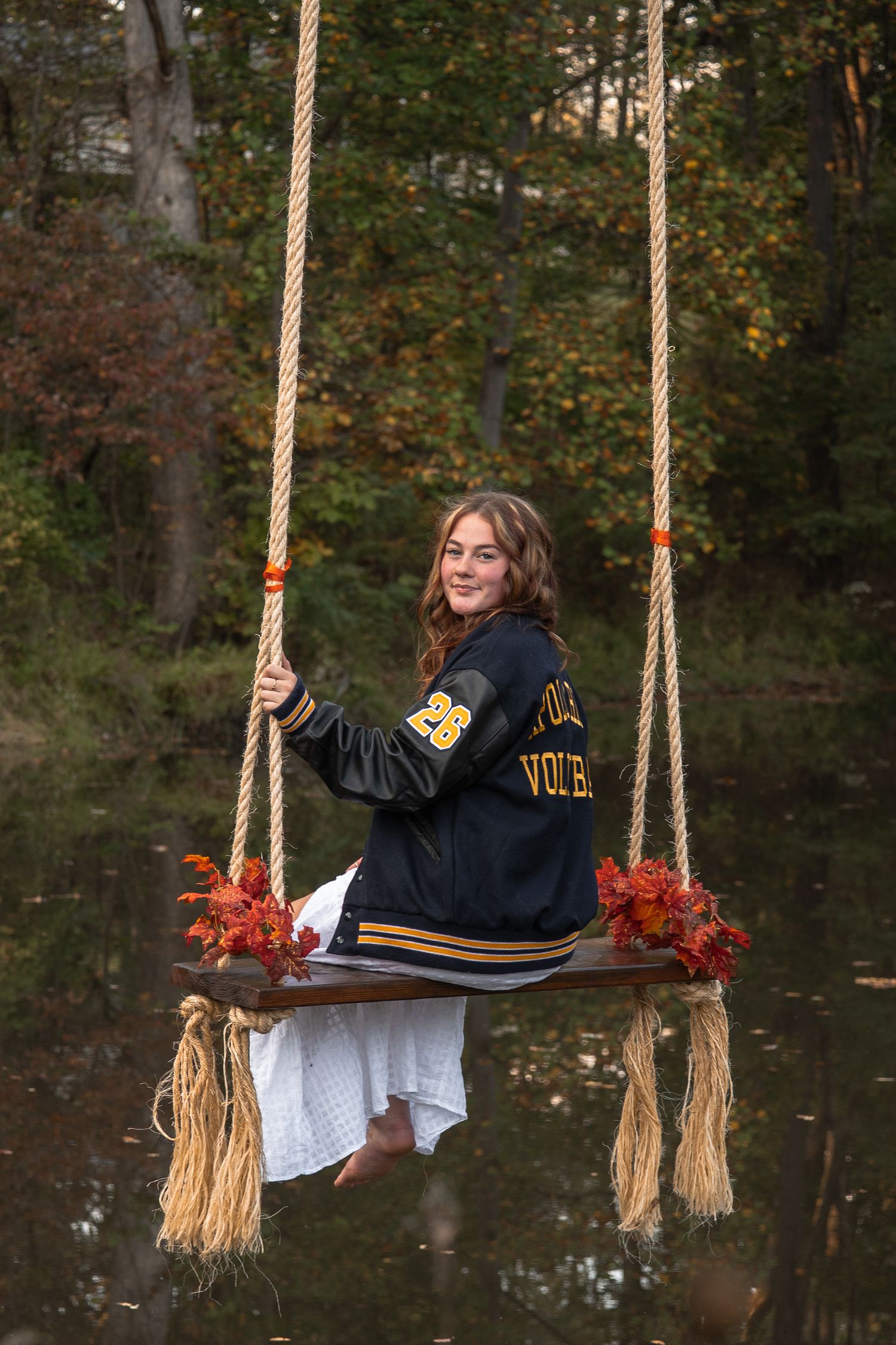 Person in a varsity jacket sits on a rustic wooden swing decorated with autumn leaves in a peaceful woodland setting.