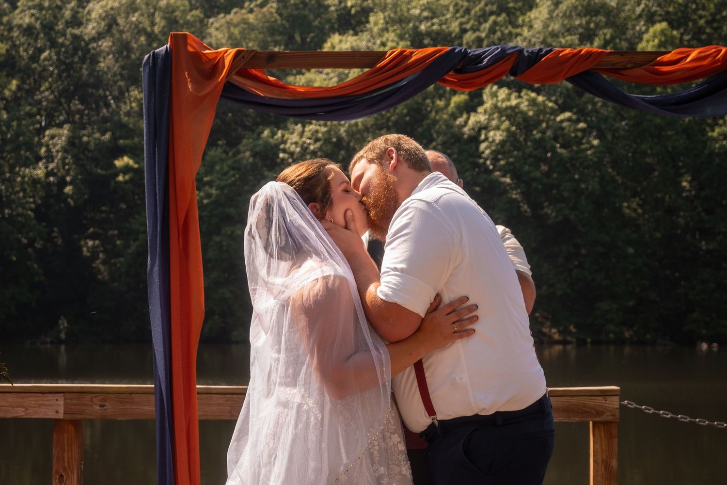 A romantic wedding couple shares intimate moments under a rustic wooden arch decorated with flowing red fabric outdoors.