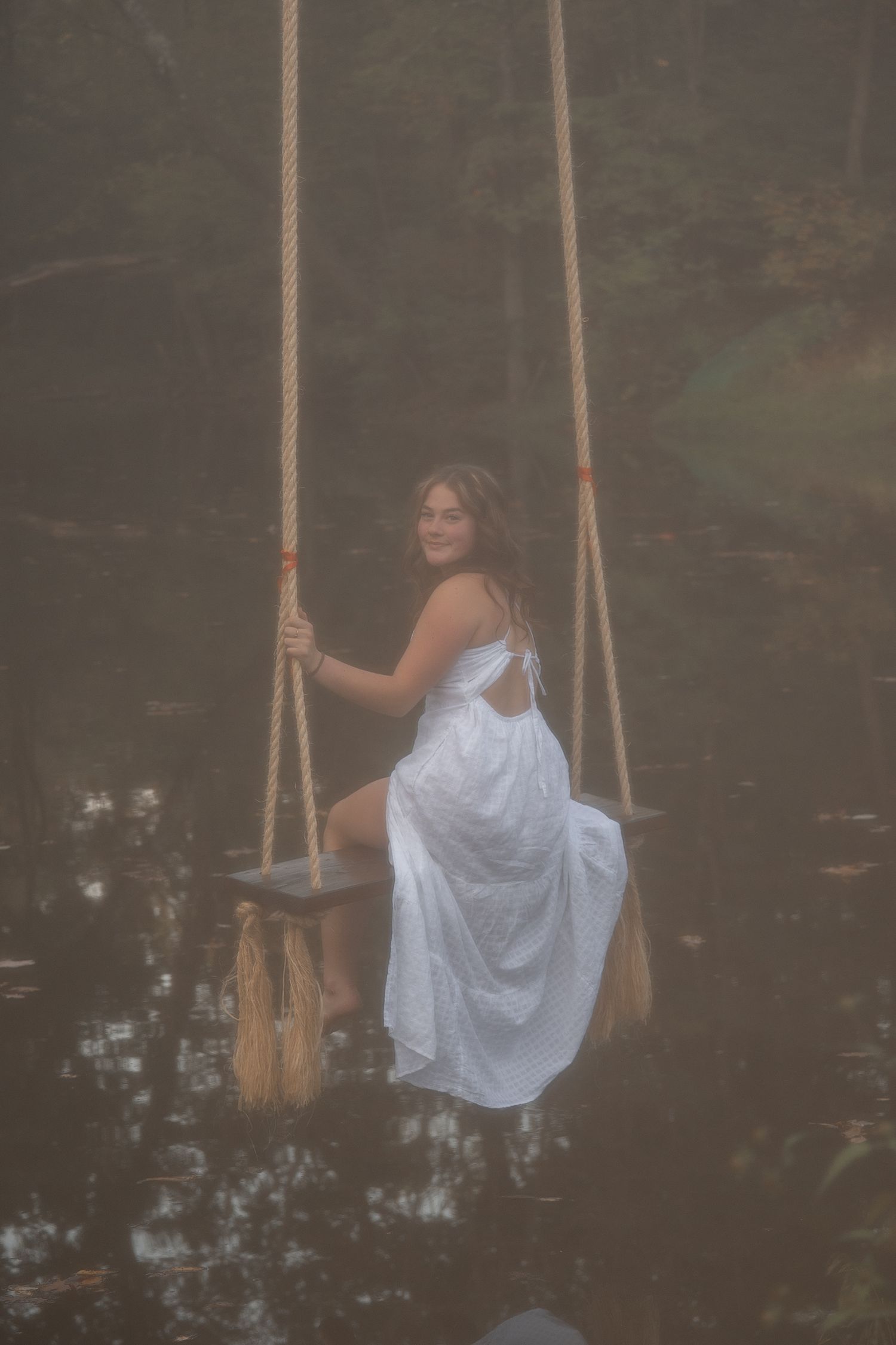 A person in a white dress swings dreamily over dark water in a moody, atmospheric forest setting.